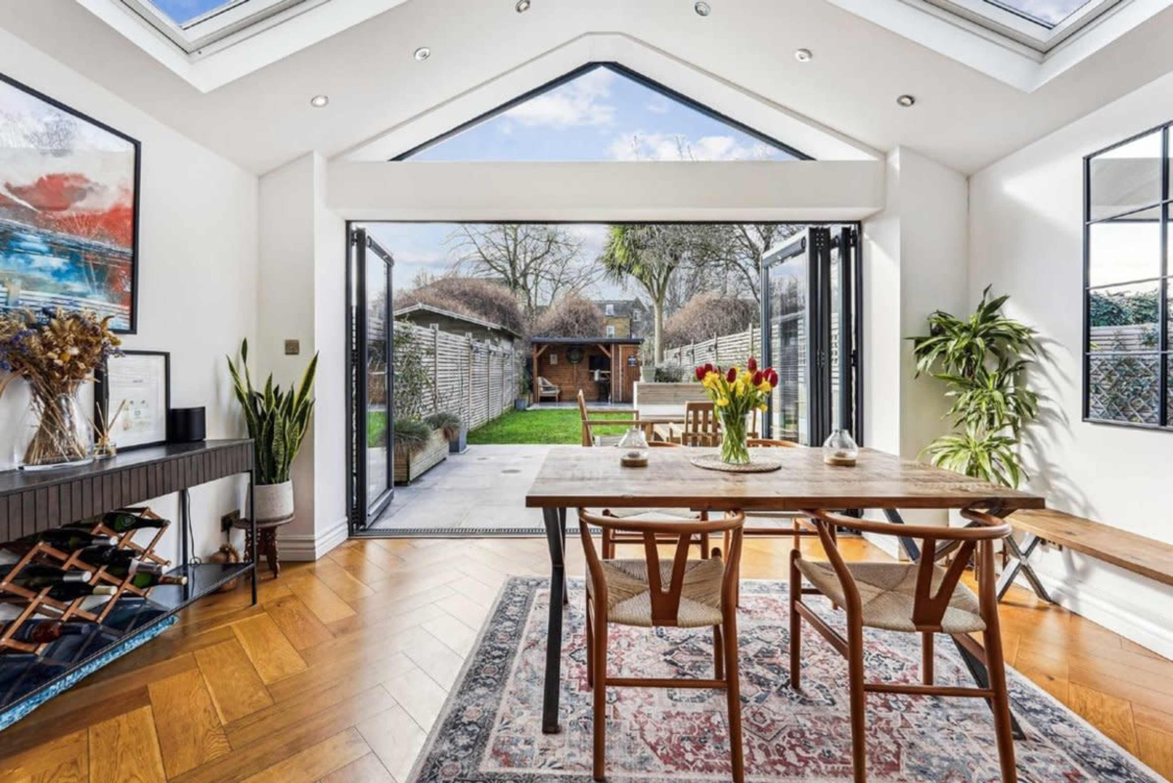 A dining area with a wooden table is illuminated by natural light from large glass doors that open to a garden.