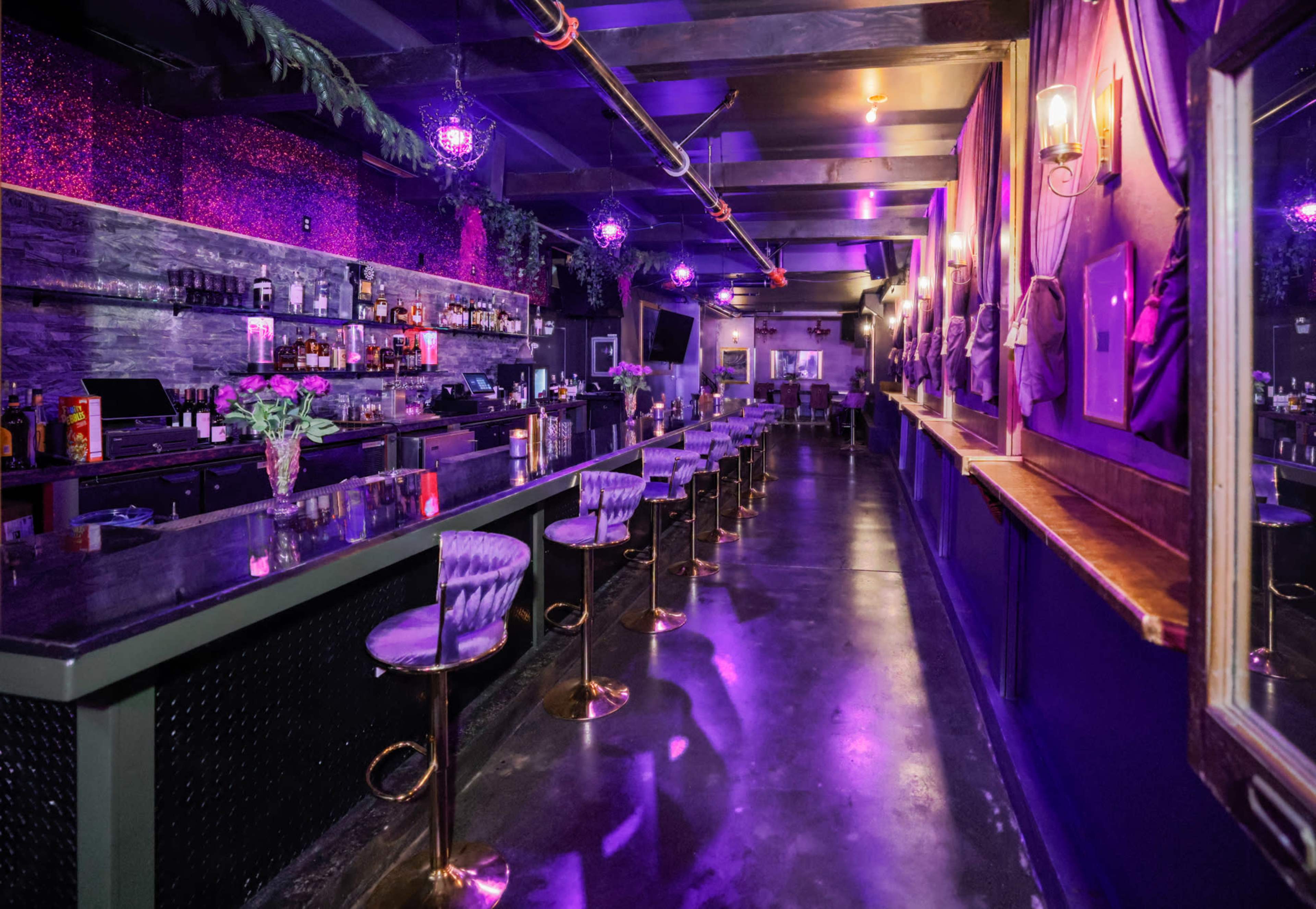 The image shows a dimly lit bar with a long counter, high stools, a selection of bottles on shelves, and purple lighting illuminating the space.