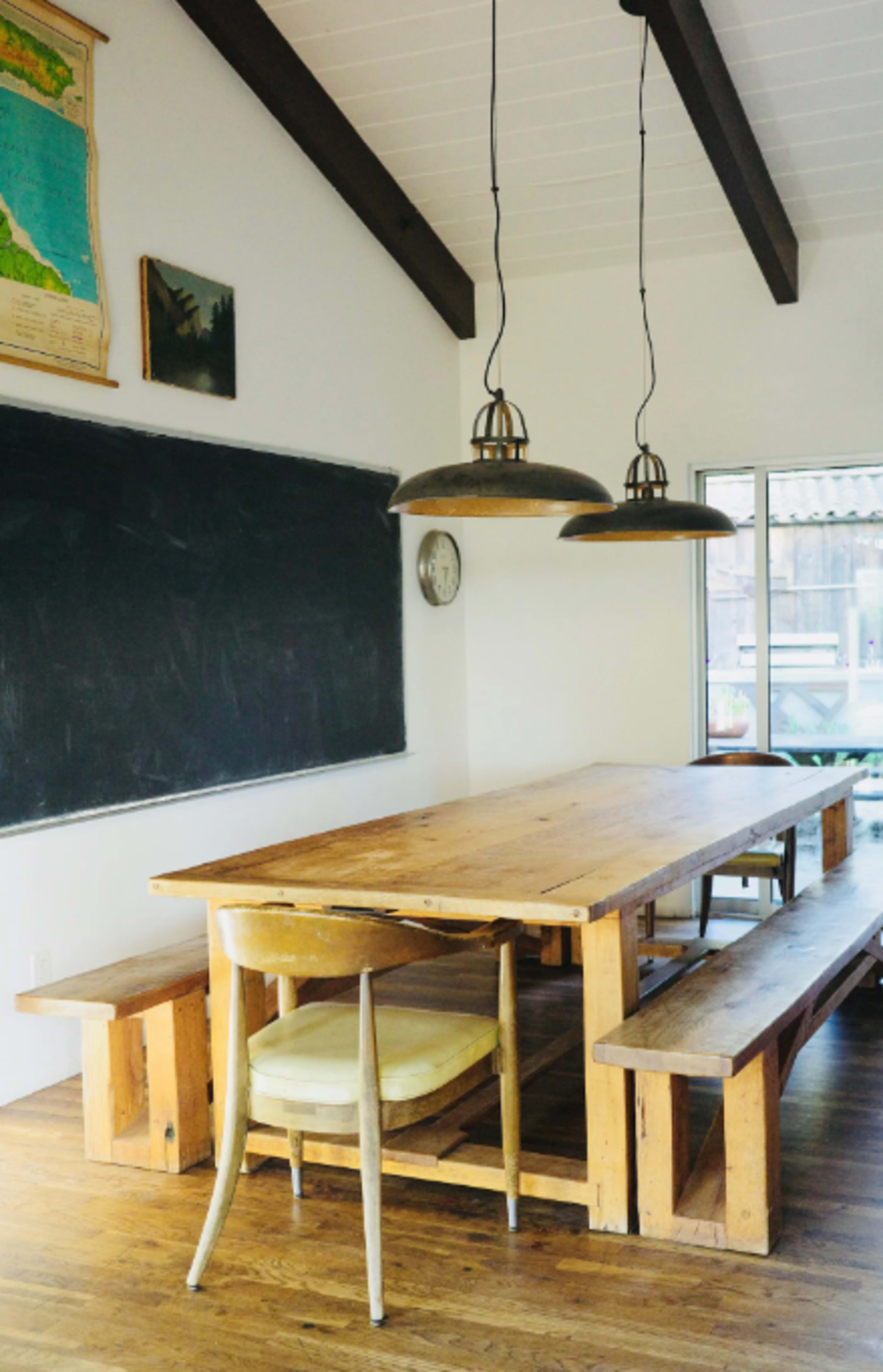 A large wooden table accompanied by benches and a chair in a room with a chalkboard and hanging light fixtures.