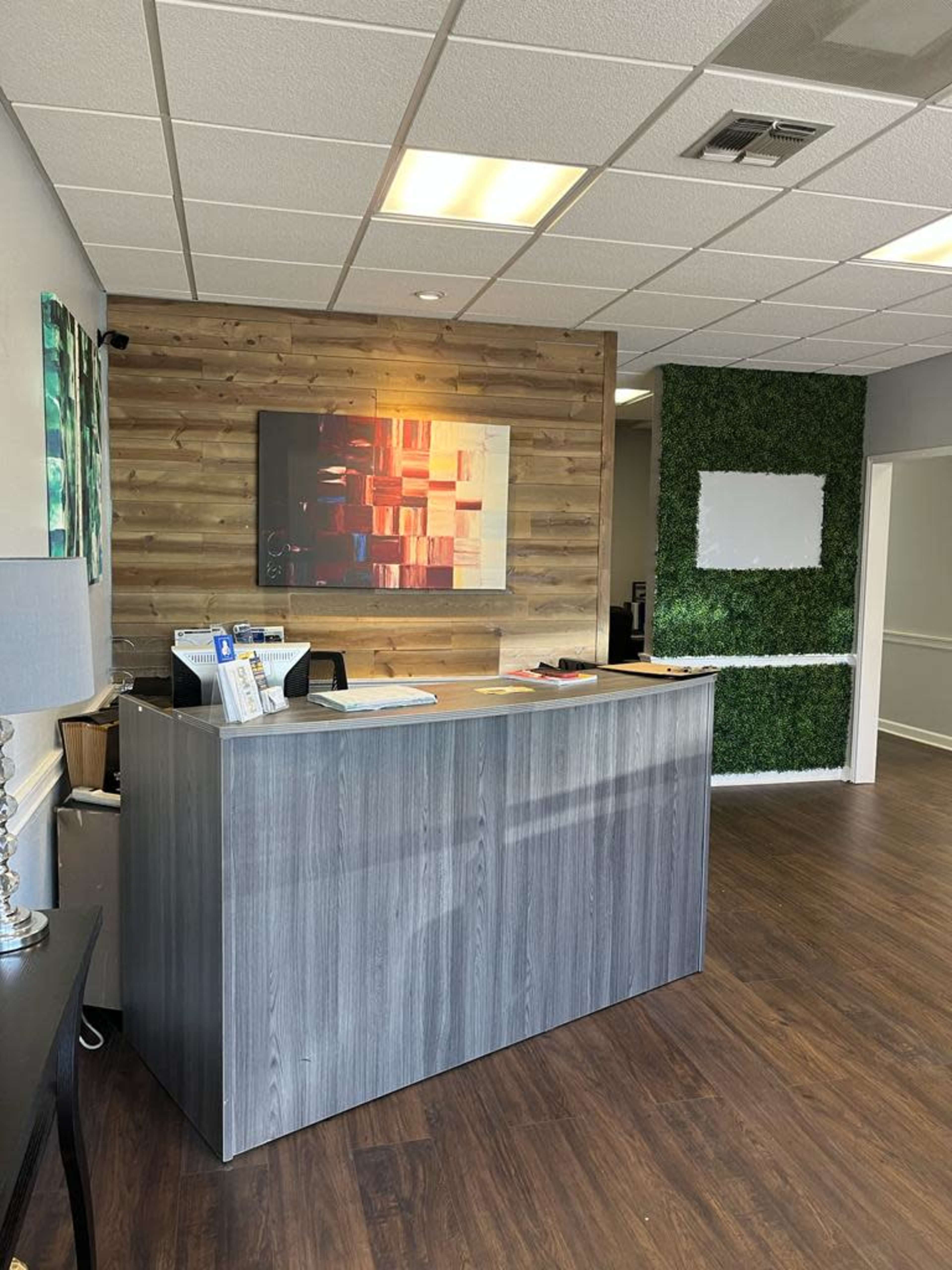 A reception area featuring a wooden front desk, a decorative painting on the wall, and a green plant wall in the background.