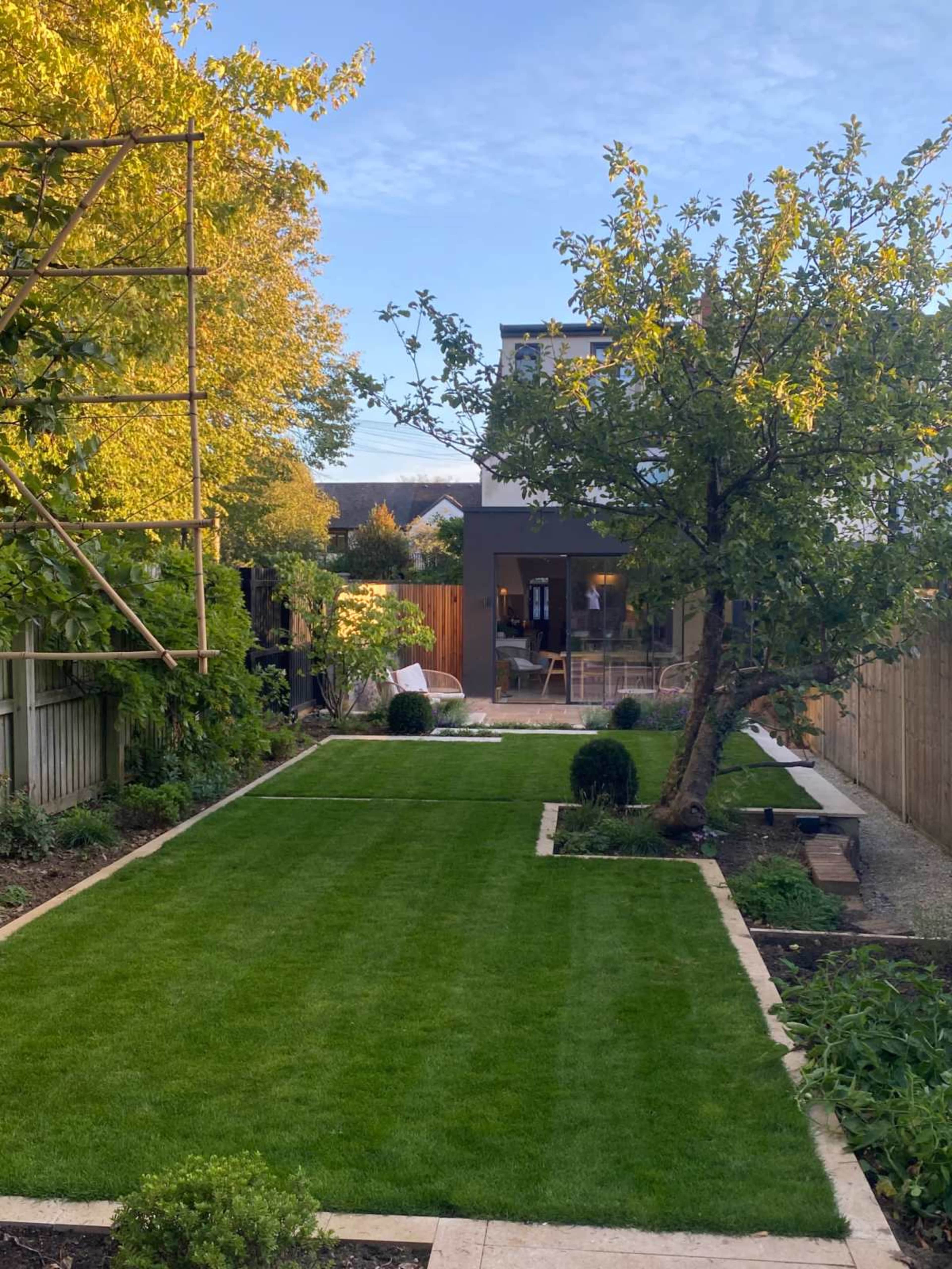 The image shows a well-maintained lawn with a central path, bordered by trees and shrubs, leading to a modern house in the background.
