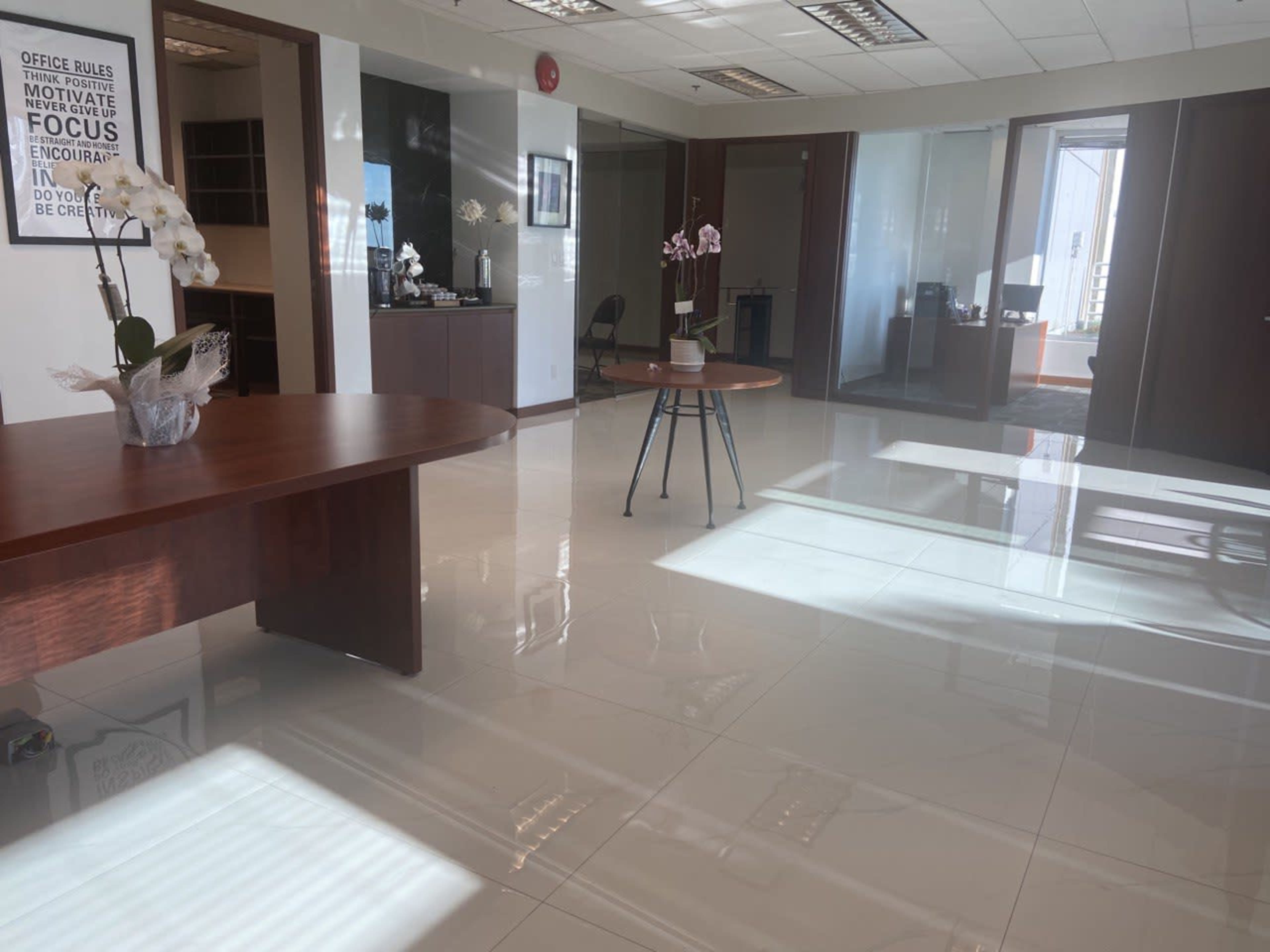 A modern office space with two round wooden tables, white tiled floors, and a large window allowing natural light to pass through.