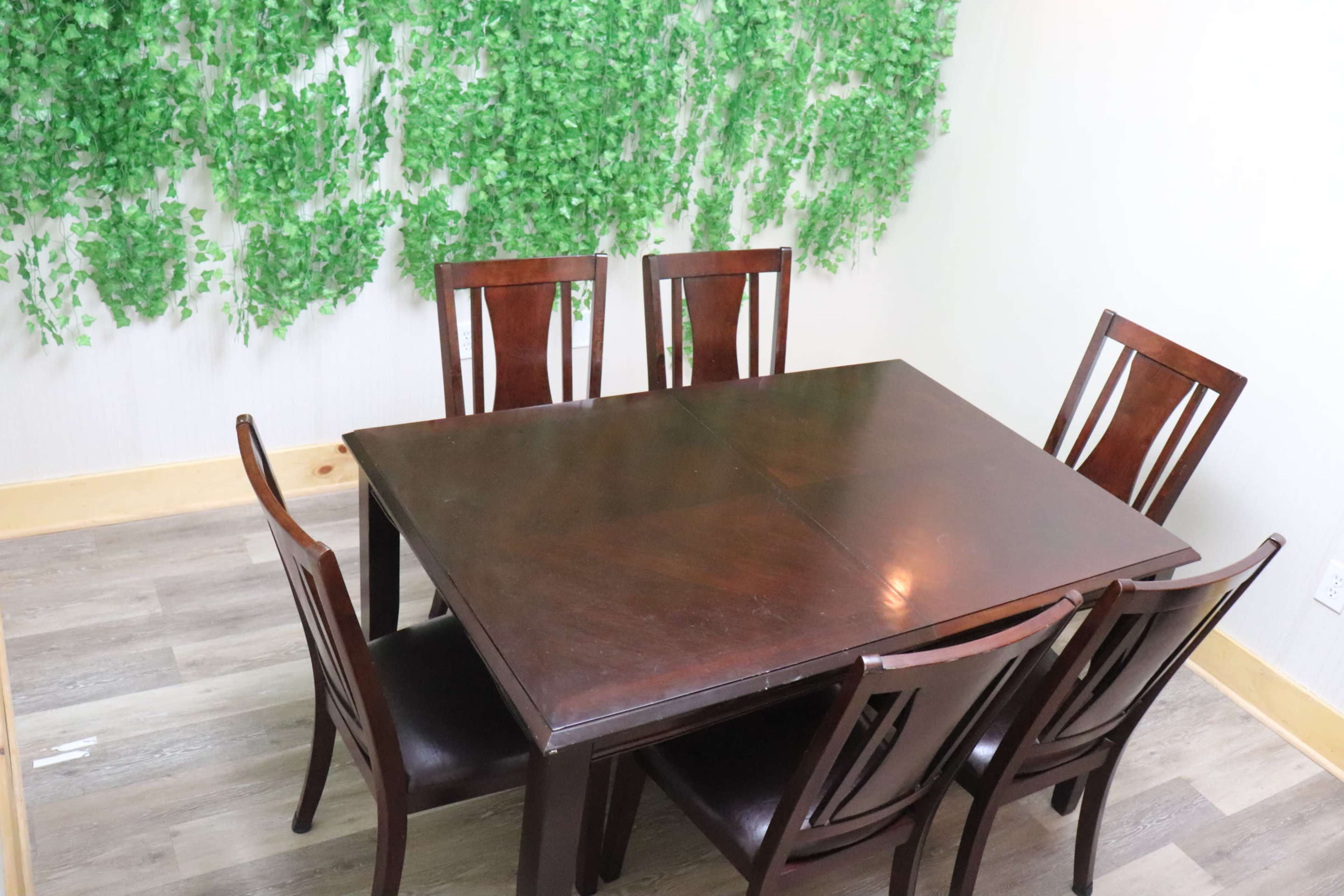 A dark wooden dining table with six chairs is set against a wall covered in artificial green vines.
