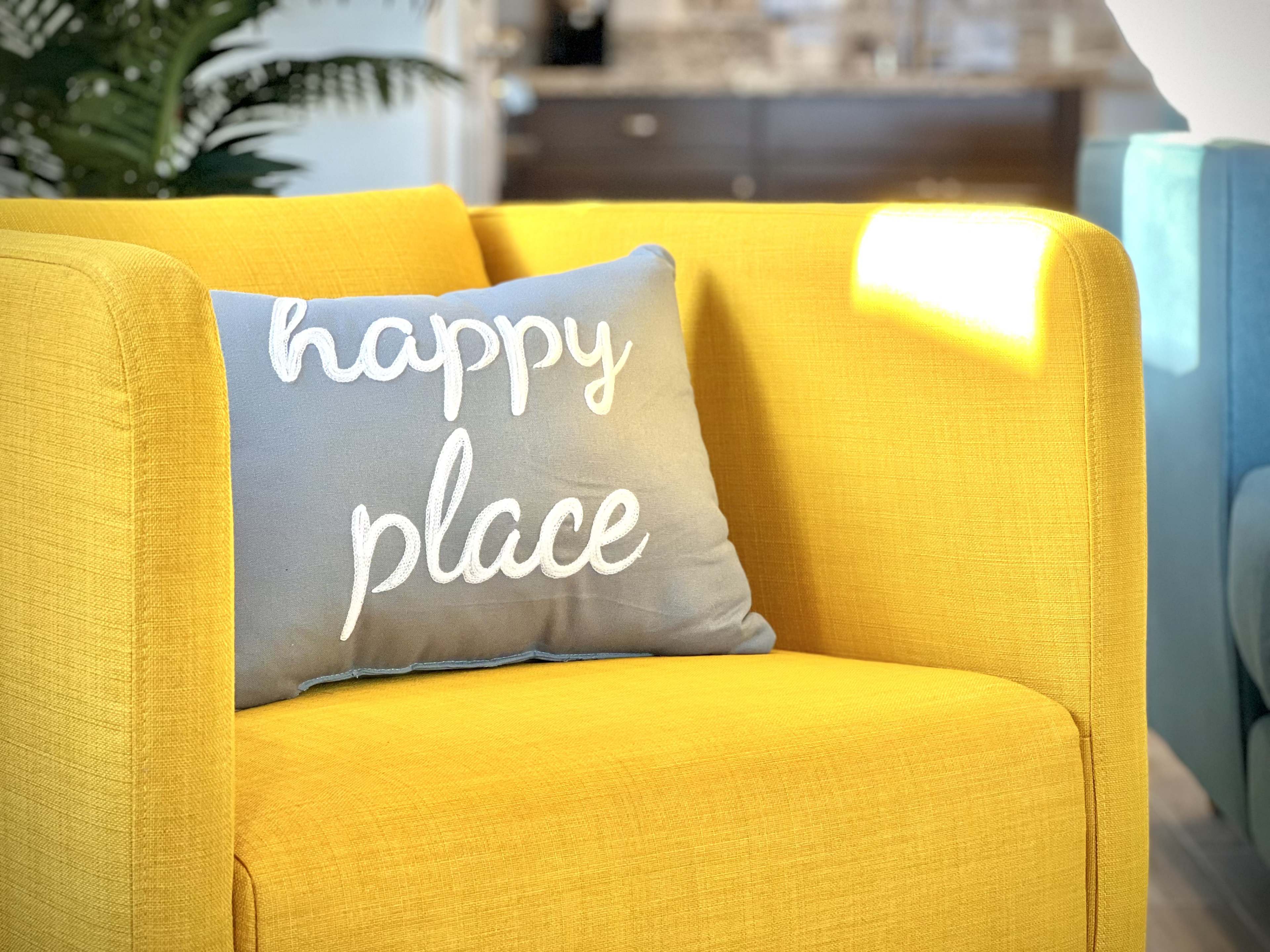 A yellow couch with a blue throw pillow that reads "happy place" rests in a well-lit room.