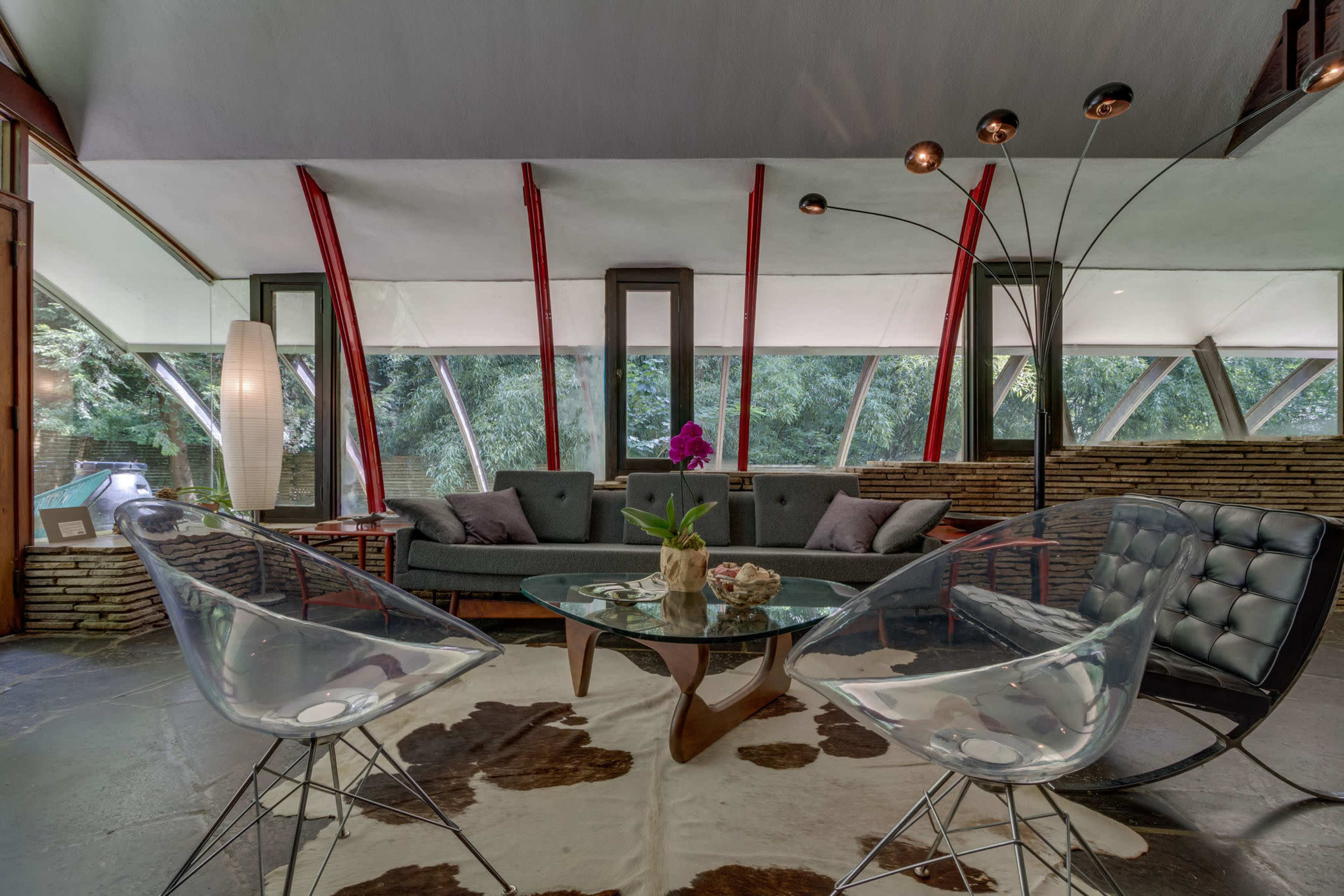 A modern living room with large windows, two clear acrylic chairs, a glass coffee table, and a sectional sofa, all set against a stone wall and surrounded by greenery.