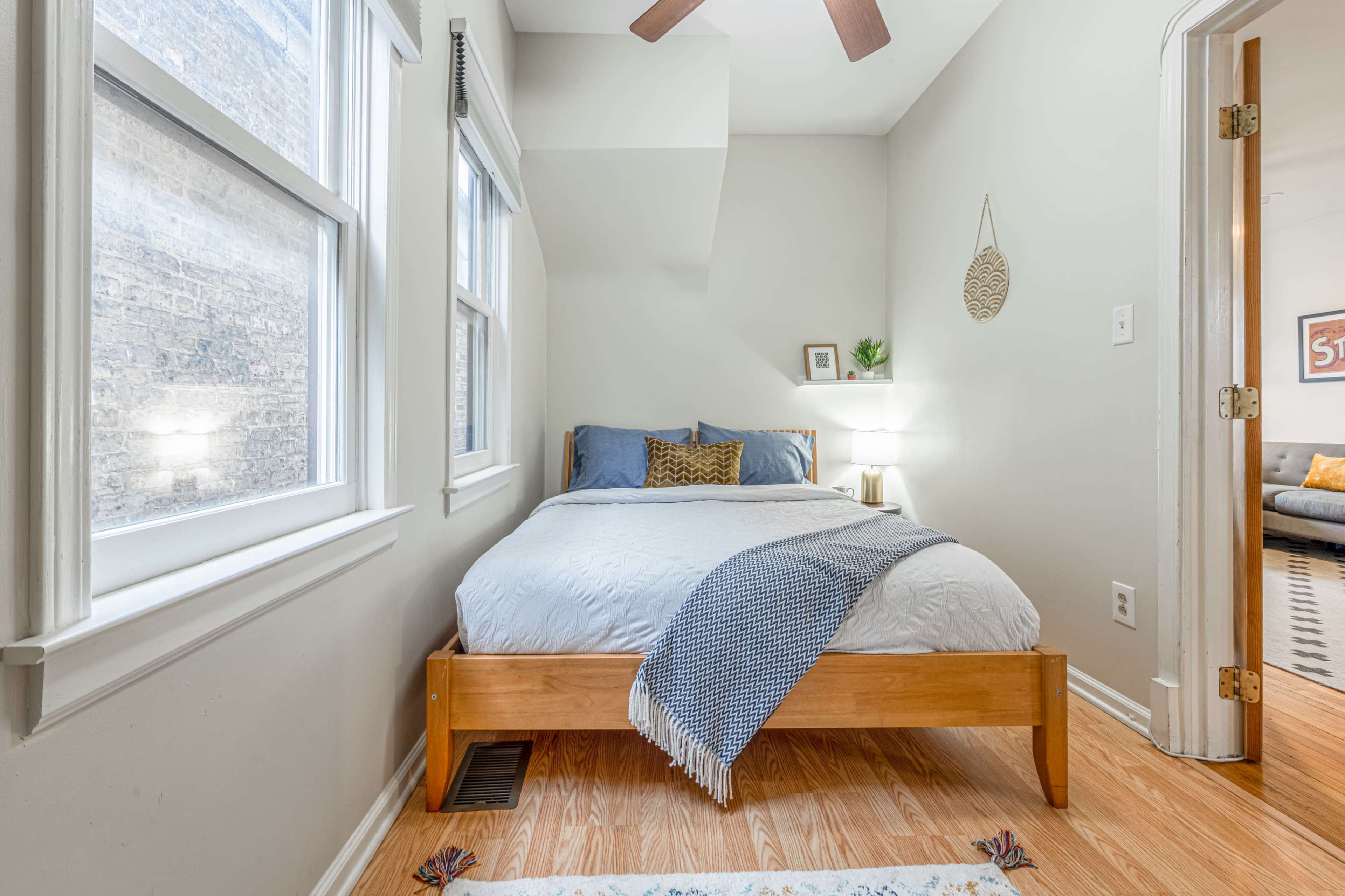 The image shows a small, bright bedroom featuring a wooden bed with white bedding, a cozy blanket, and natural light coming through two windows.