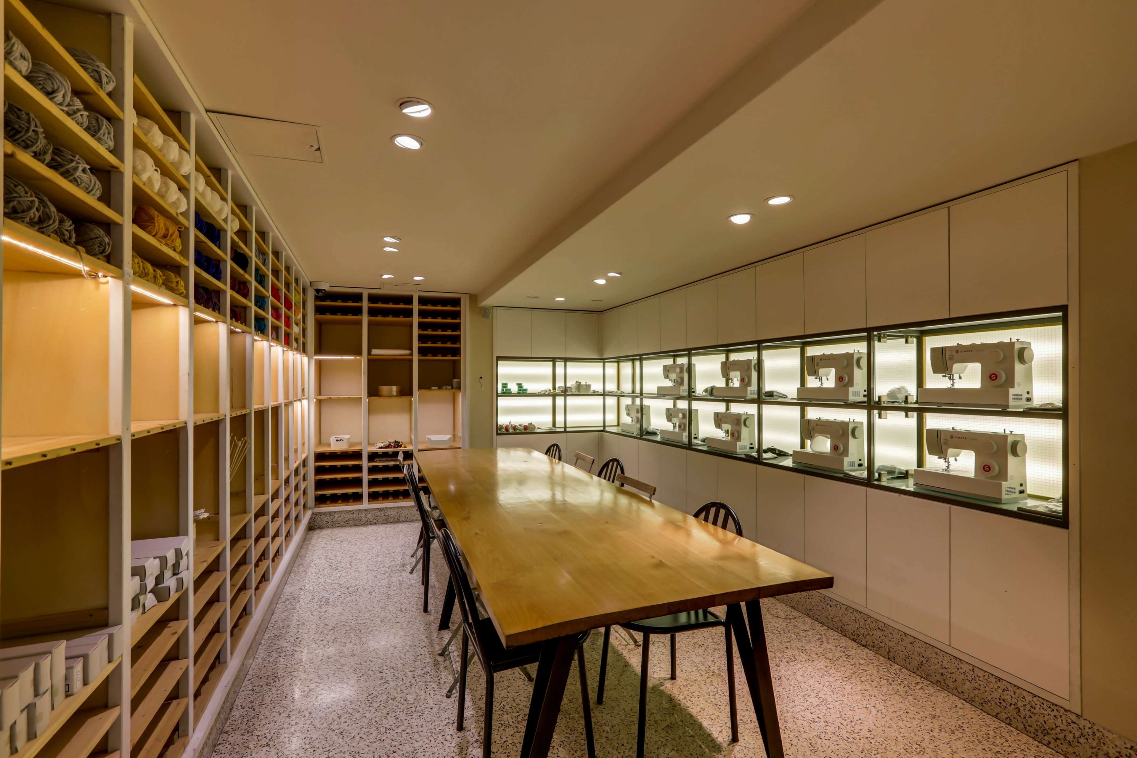 The image shows a well-organized sewing studio with a long wooden table, shelves filled with fabric rolls, and display cases containing sewing machines.