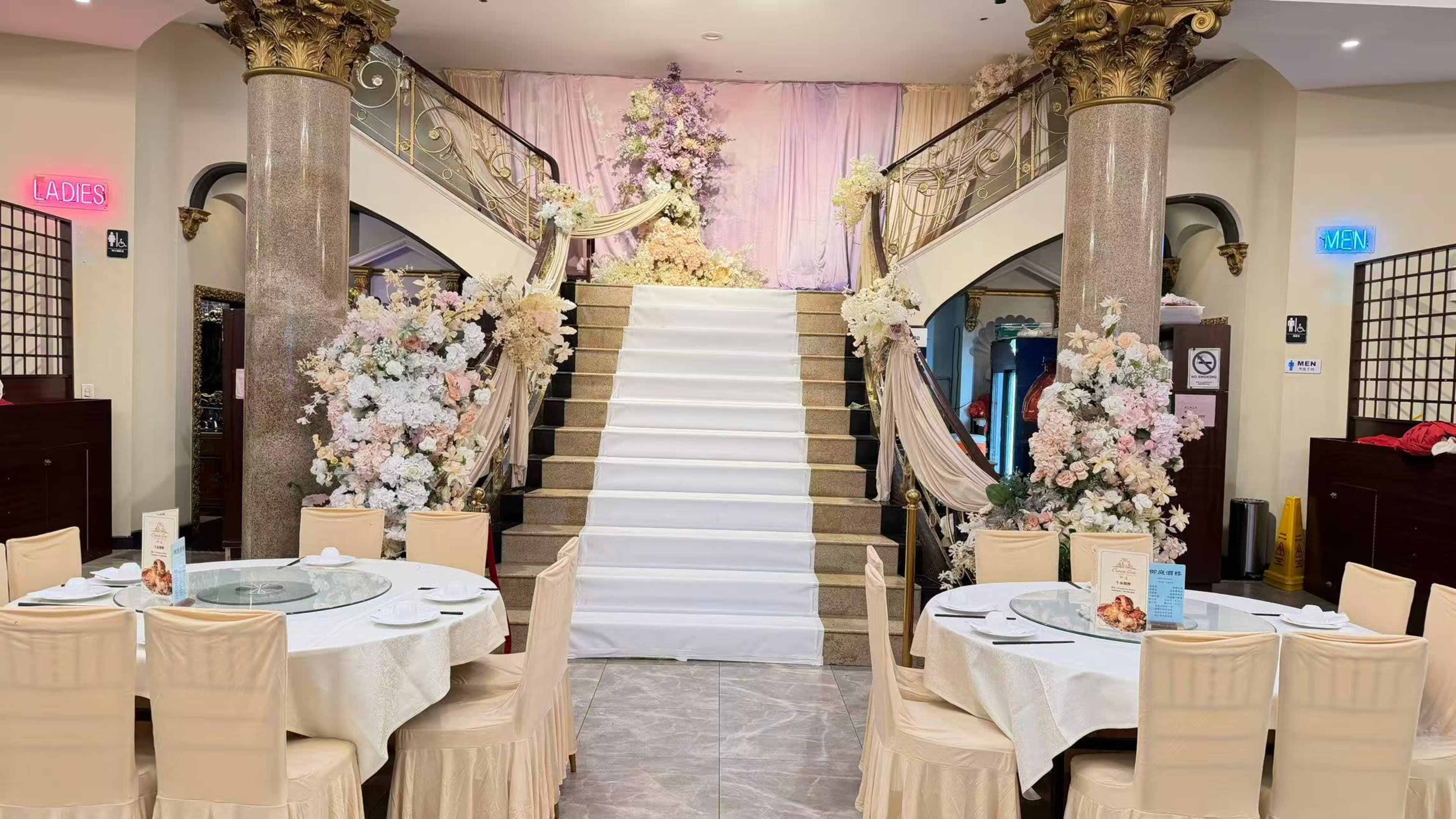 The image shows a banquet hall featuring a grand staircase flanked by floral decorations, with tables set for dining in the foreground.