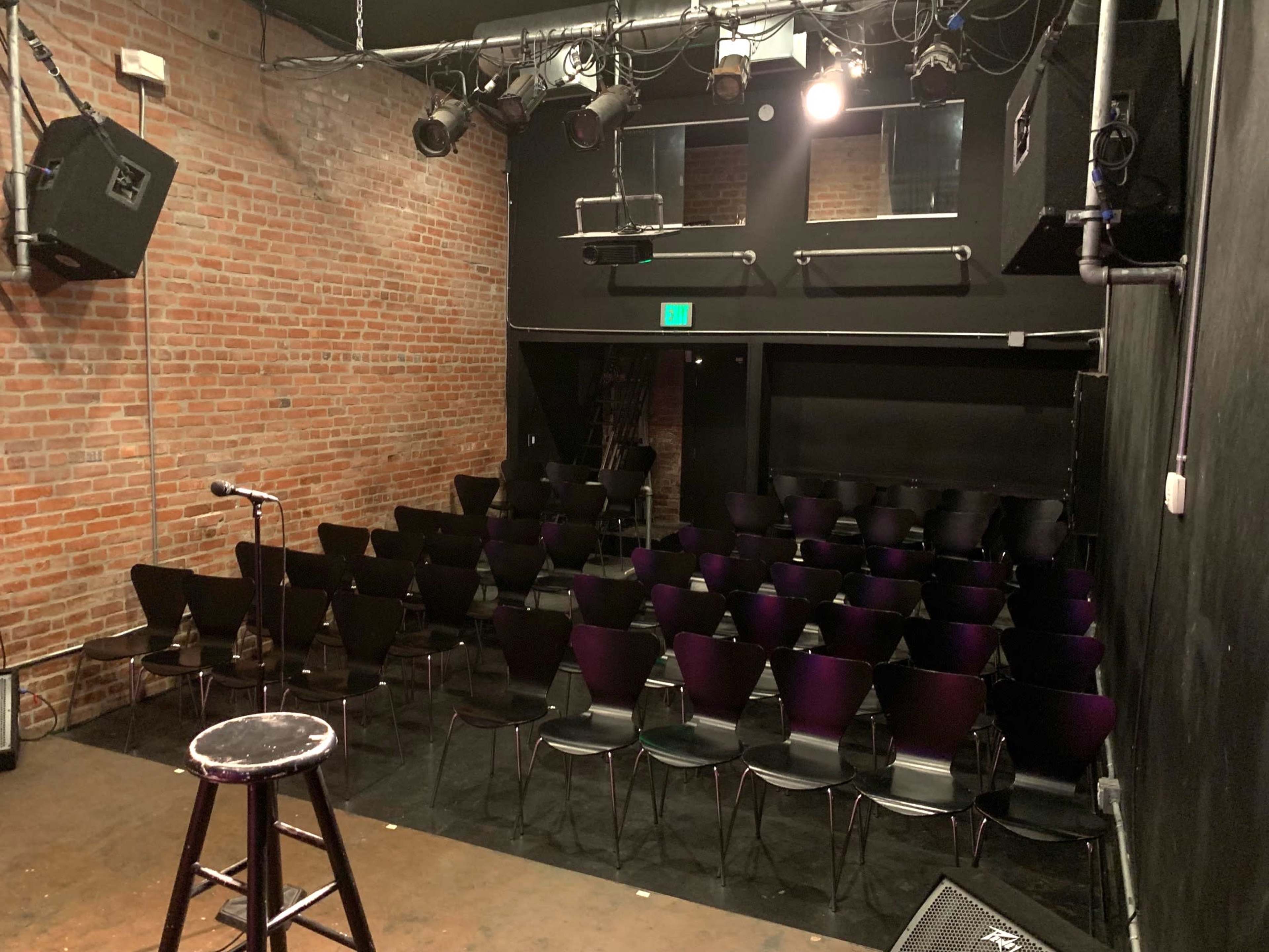 An empty comedy club stage with a microphone stand and a stool, facing rows of black chairs arranged for an audience against a brick wall.