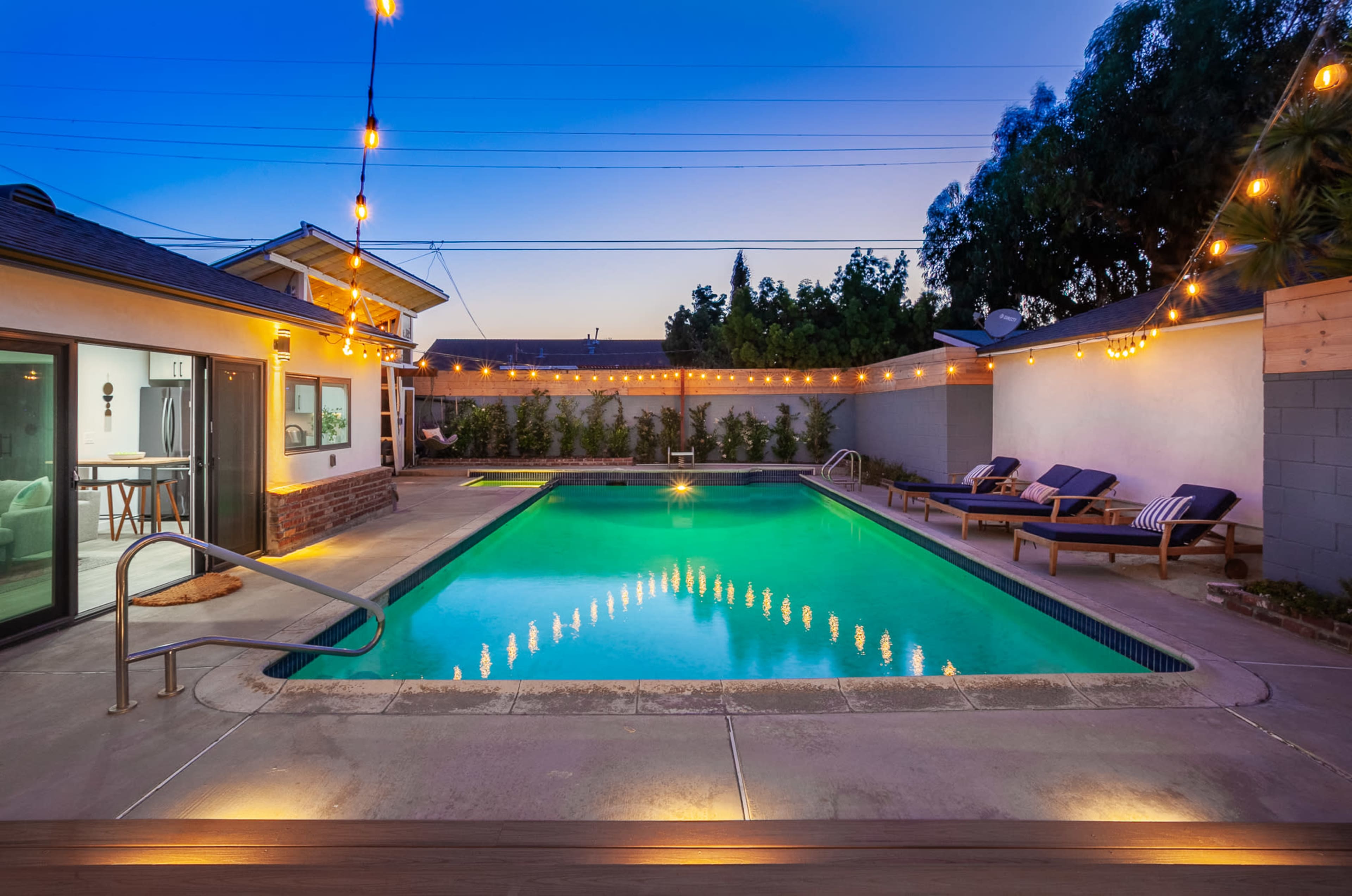 A backyard features a swimming pool surrounded by lounge chairs and string lights illuminating the area at dusk.