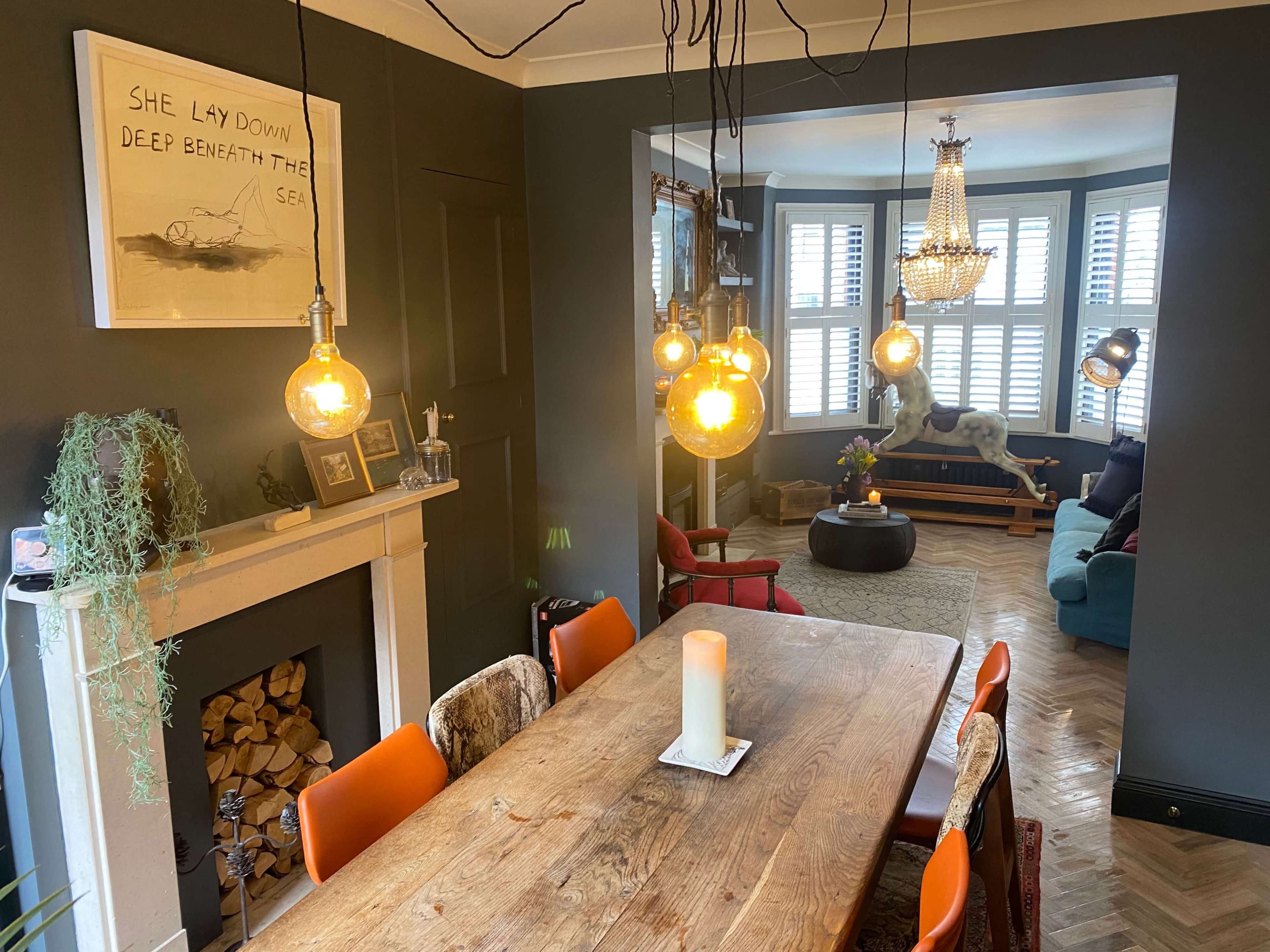A dining area with a wooden table and orange chairs, featuring pendant lights above and a fireplace, leading into a cozy living room with a chandelier and a blue sofa.