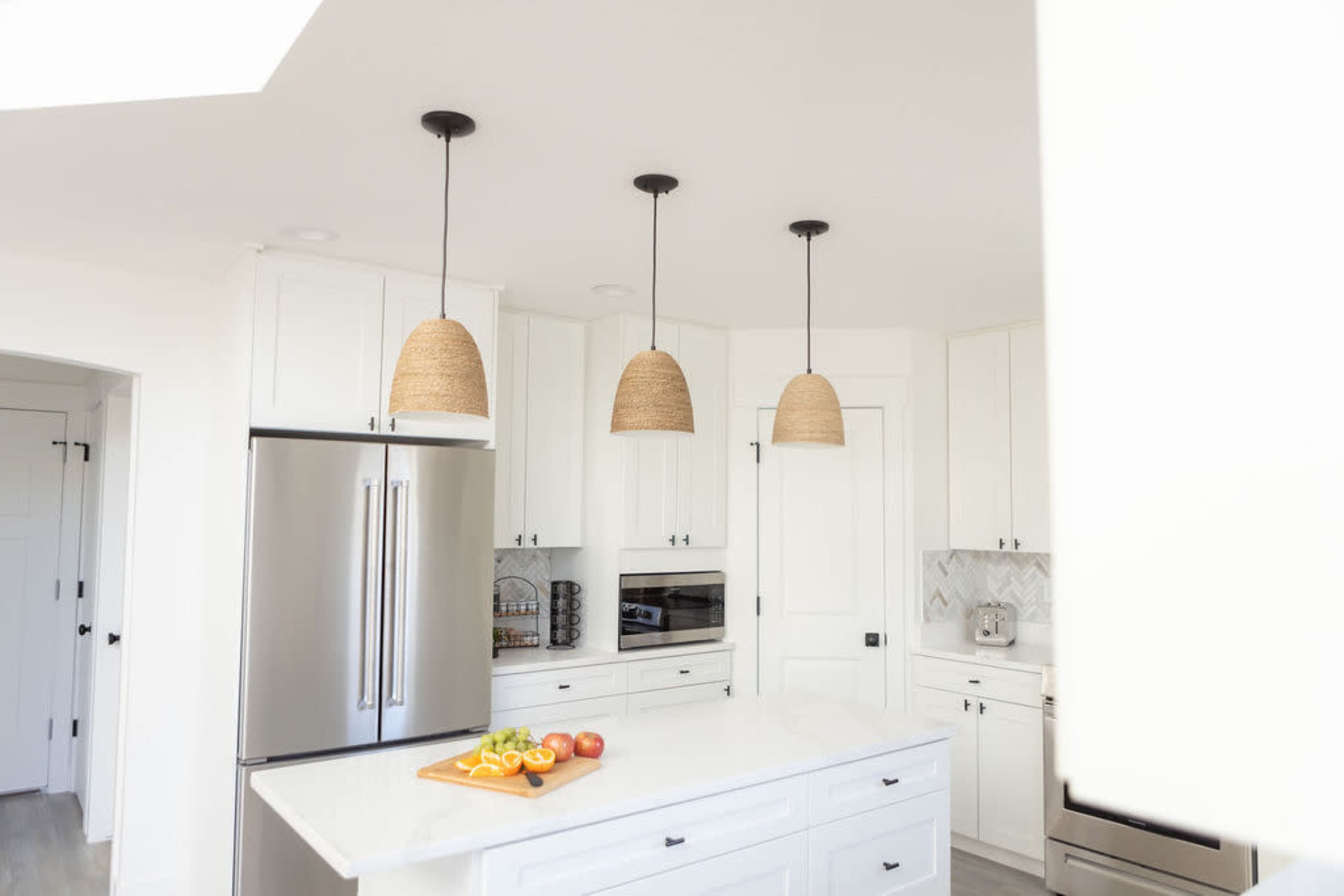 A modern kitchen features white cabinetry, stainless steel appliances, and three woven pendant lights hanging over an island with cut fruits.