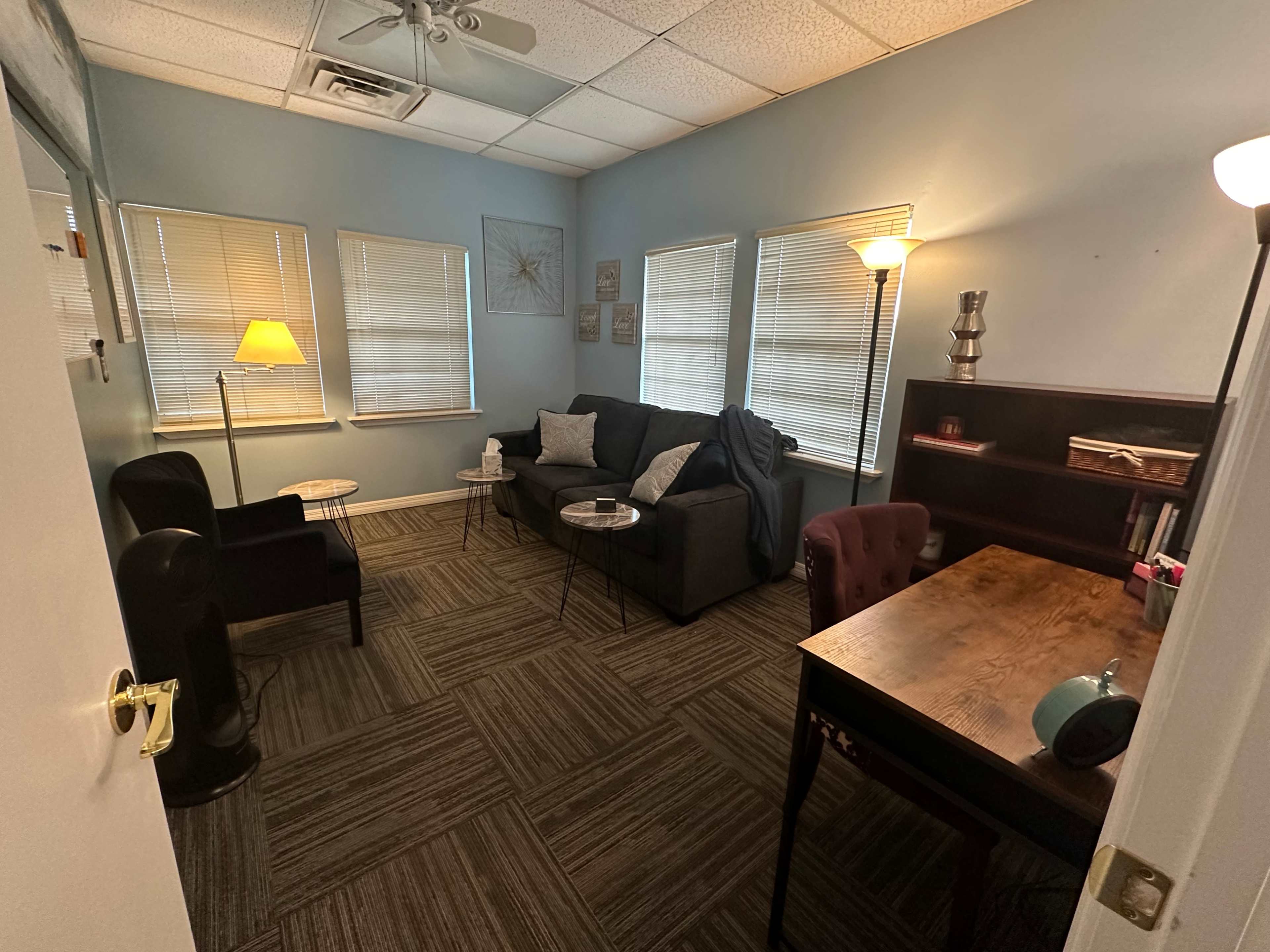 The image shows a small office space with a couch, two chairs, a coffee table, a desk, and multiple windows.