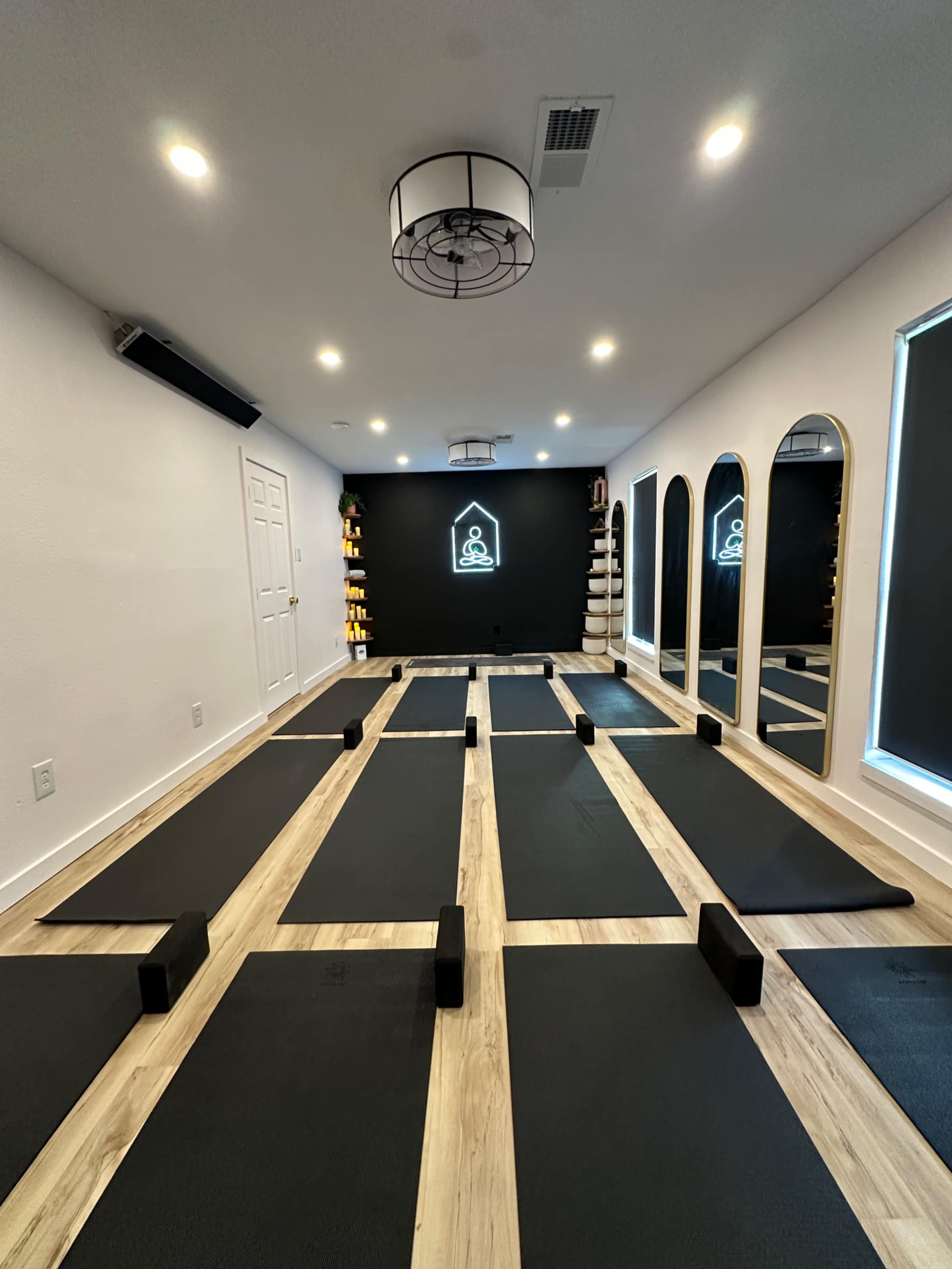 The image shows a yoga studio with several yoga mats arranged on a wooden floor, a black accent wall featuring a neon yoga logo, and mirrors on either side.