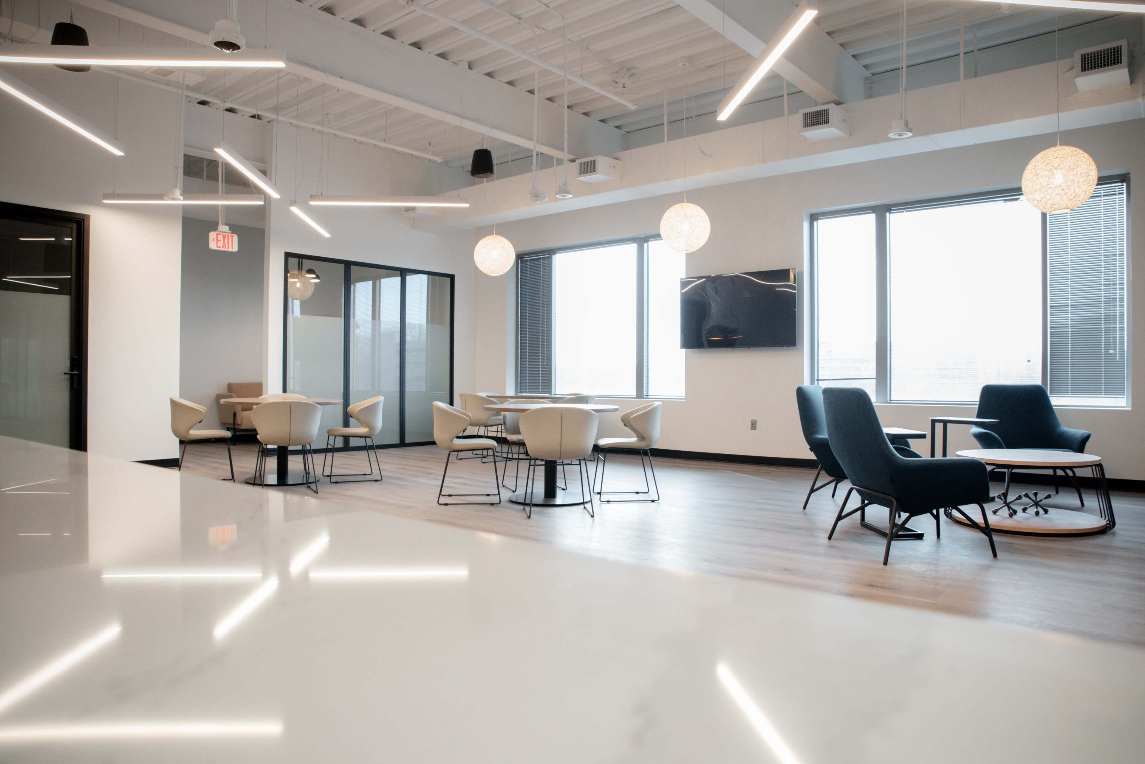 The image shows a modern office lounge area with contemporary seating, round tables, and large windows providing natural light.