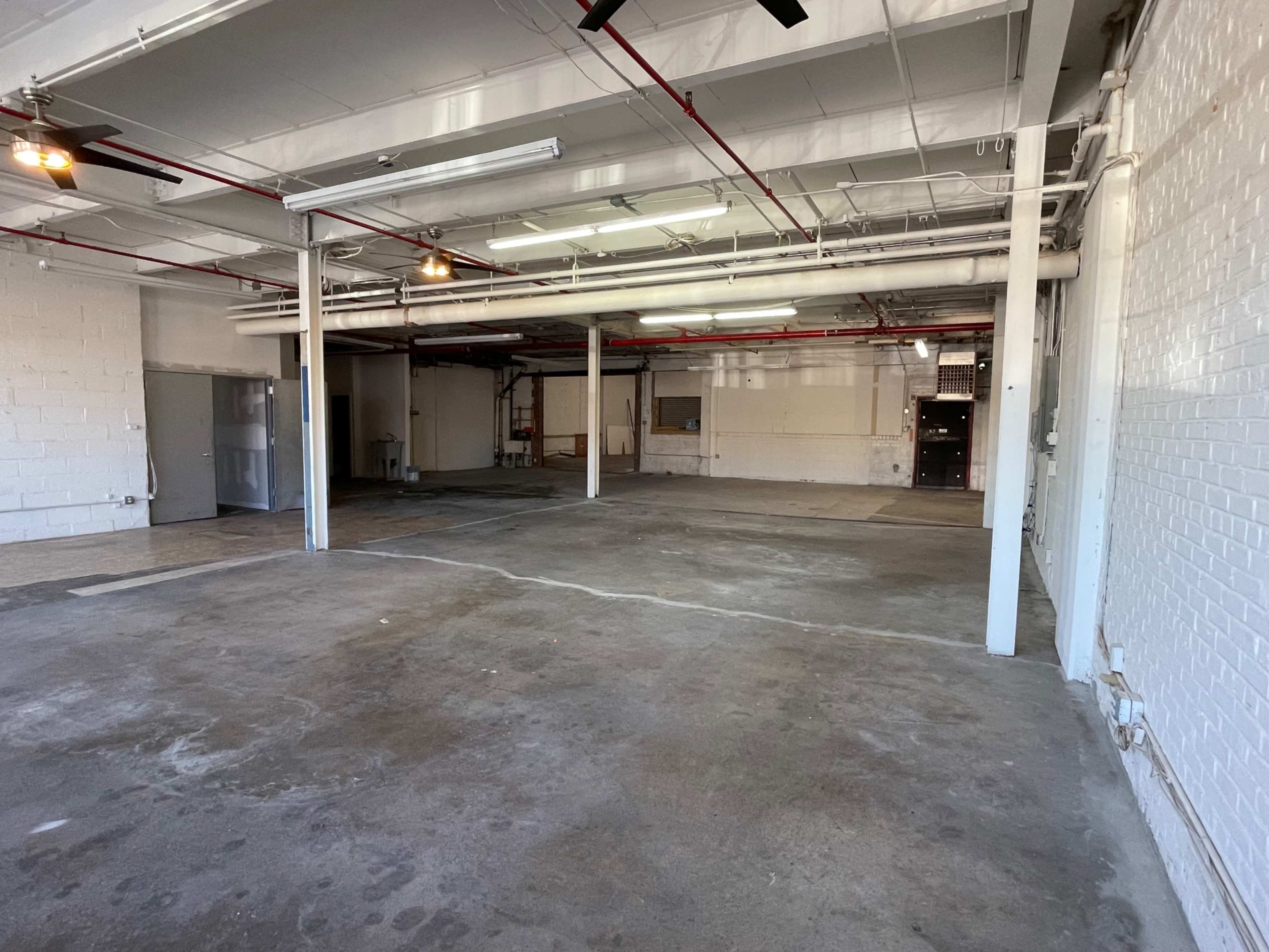 The image shows an empty industrial space with concrete flooring, exposed pipes, and white brick walls.