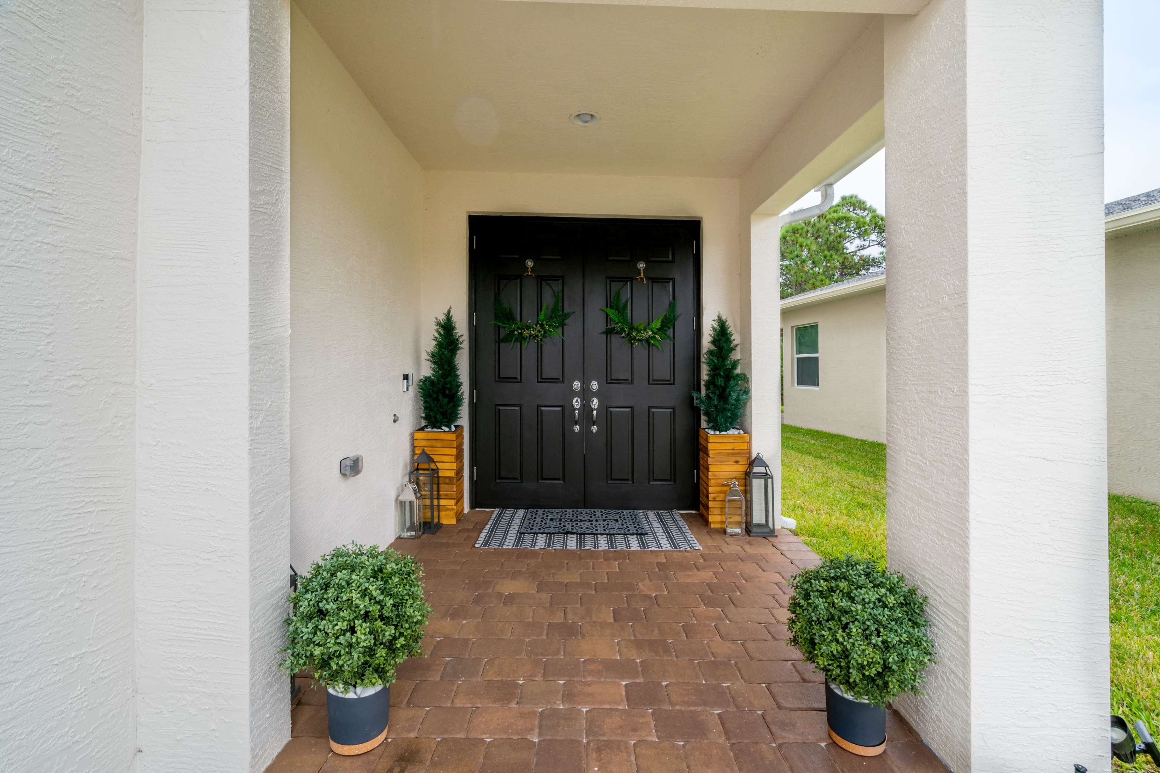 A front entrance with double black doors, flanked by two potted greenery arrangements and wooden boxes, leading to a stone pathway.