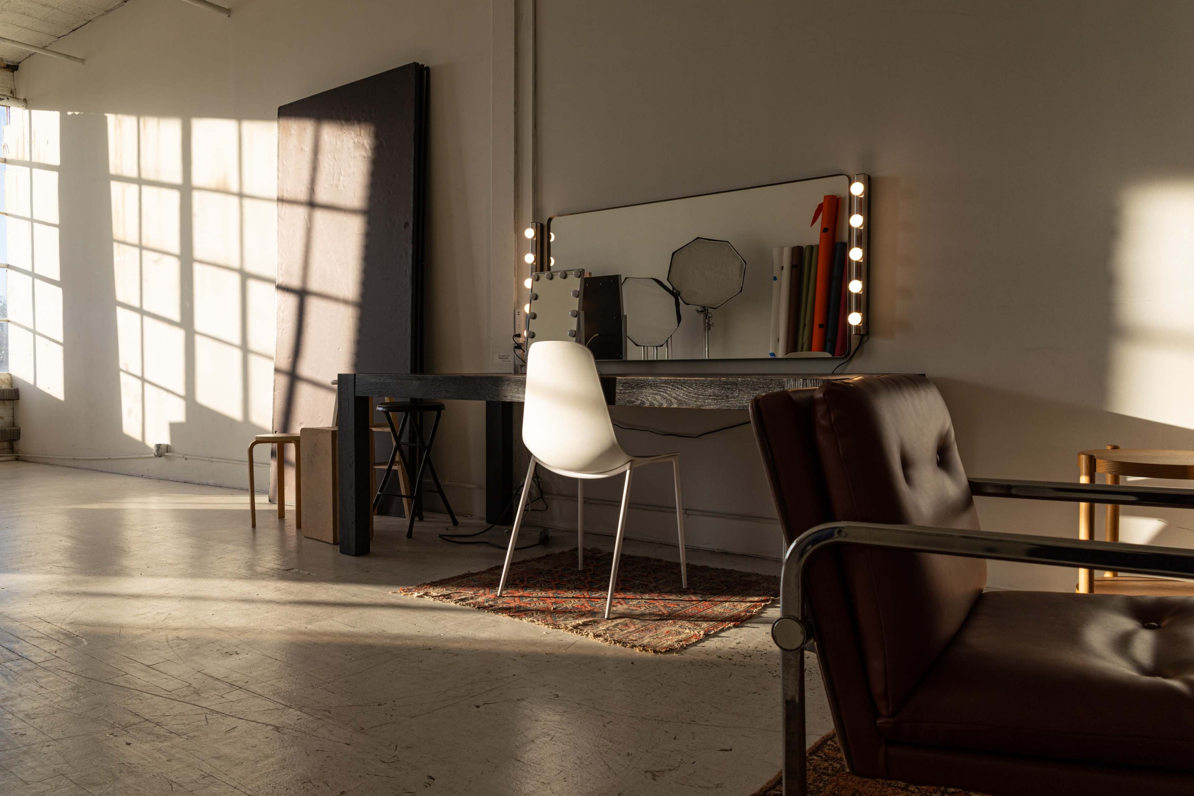 The image shows a bright room with a mirror, a simple chair, and a dressing table illuminated by light bulbs, along with a few pieces of furniture and sunlight casting shadows on the floor.