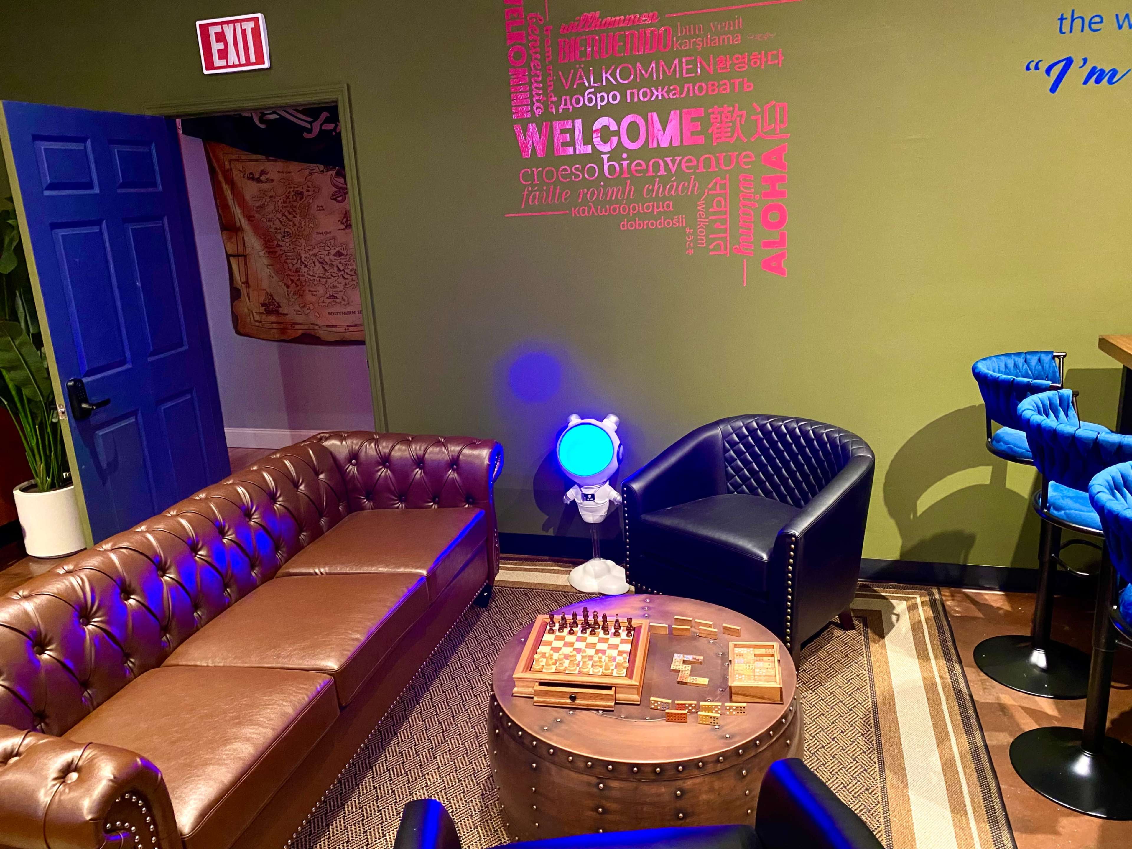 The scene features a cozy seating area with a brown leather sofa, a black armchair, a chess table, and a blue light in the corner, surrounded by walls adorned with welcome messages in various languages.