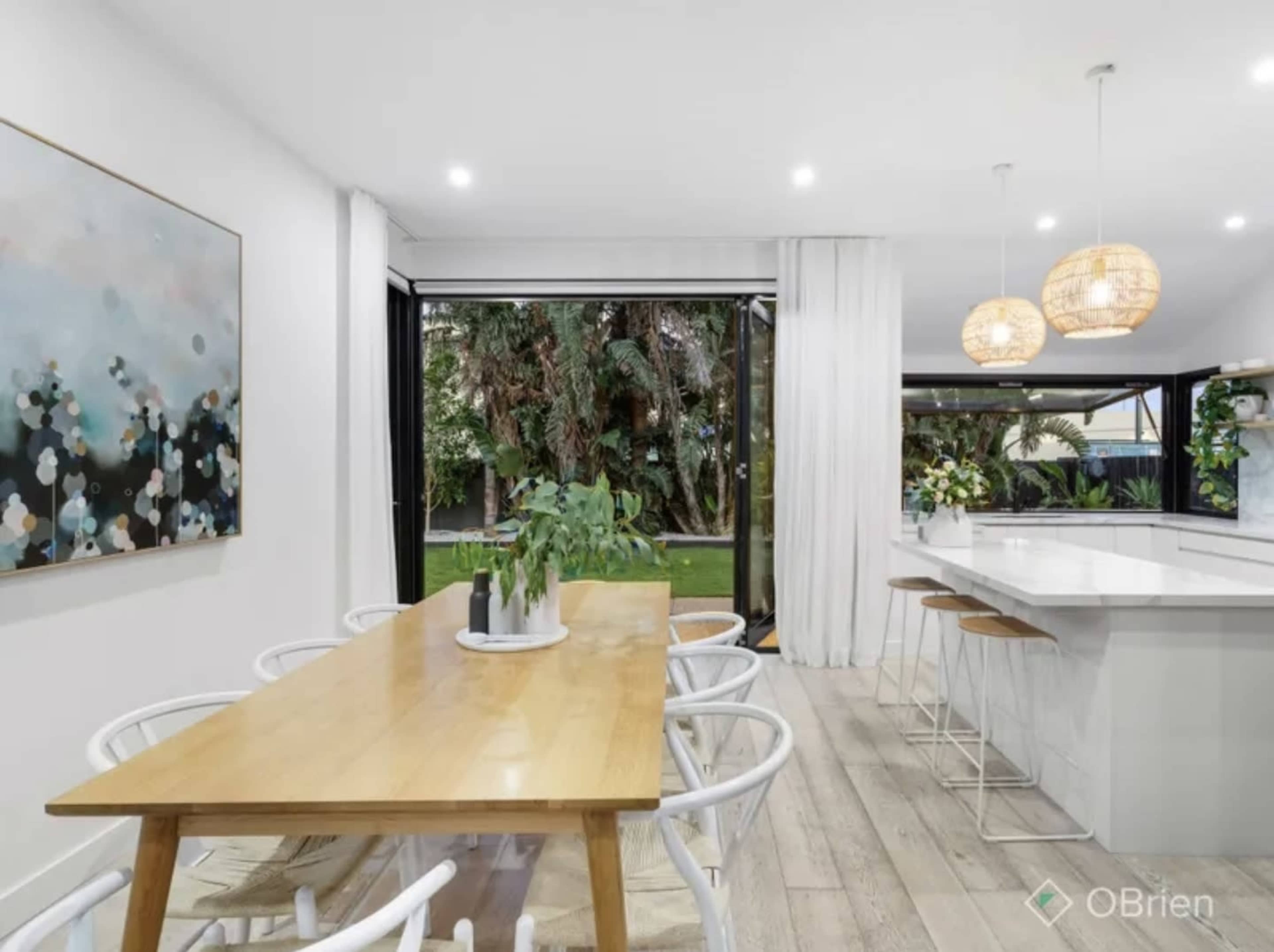 The image shows a dining area with a wooden table, white chairs, and a large painting on the wall, adjacent to a kitchen with bar stools and views of an outdoor garden.