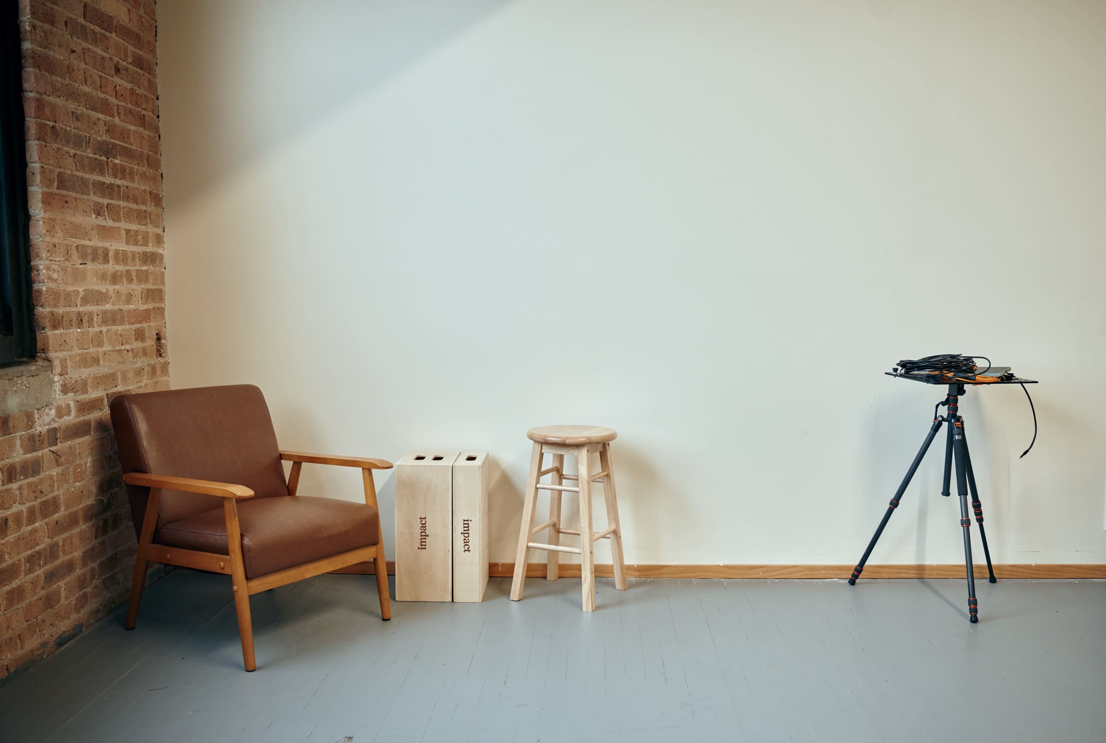 The image shows a simple interior space featuring a brown armchair, two wooden boxes labeled "material," a wooden stool, and a camera tripod.