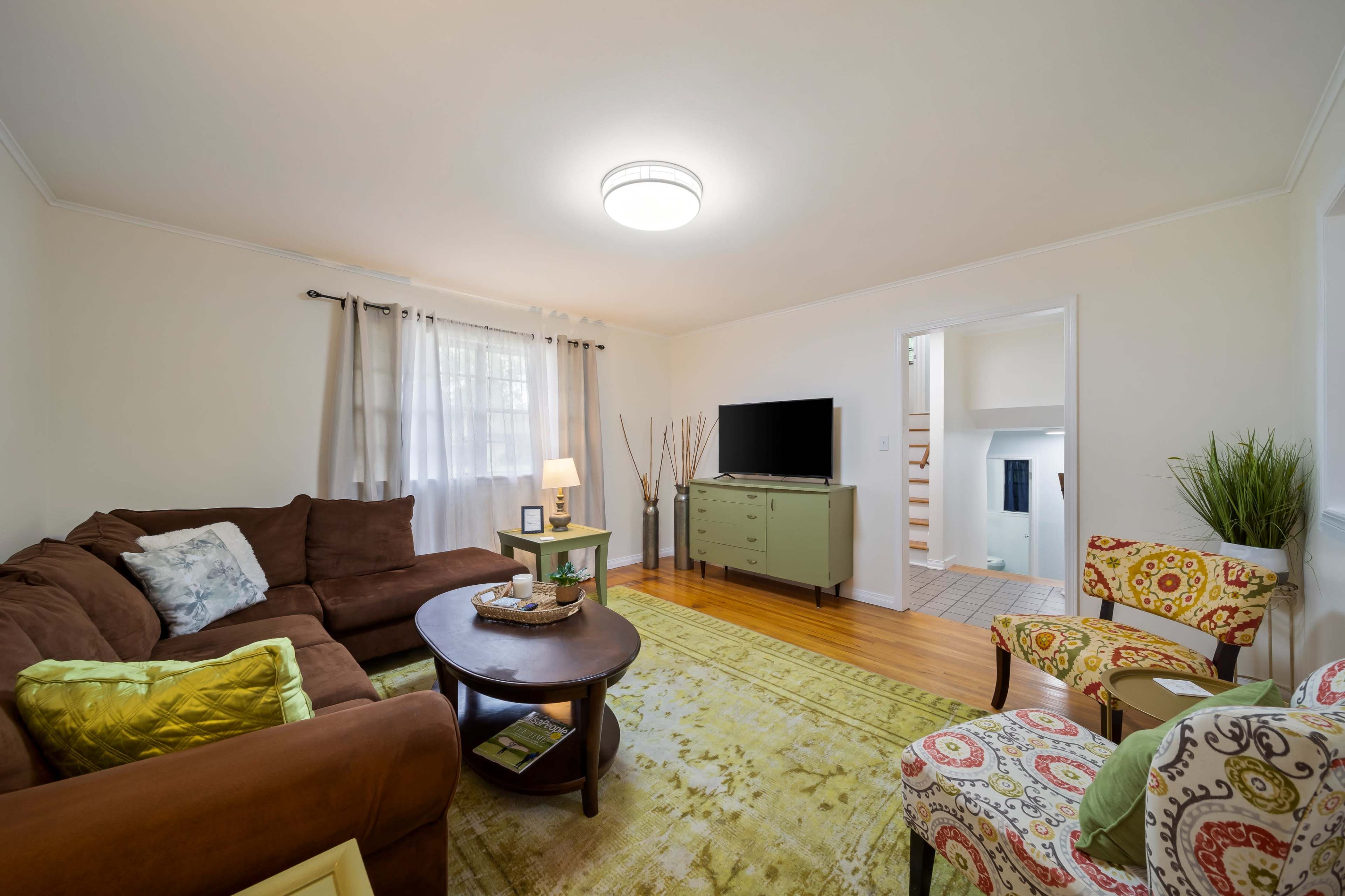 A cozy living room features a brown sectional sofa, a round coffee table, a green TV stand, and patterned chairs, with light streaming in through sheer curtains.