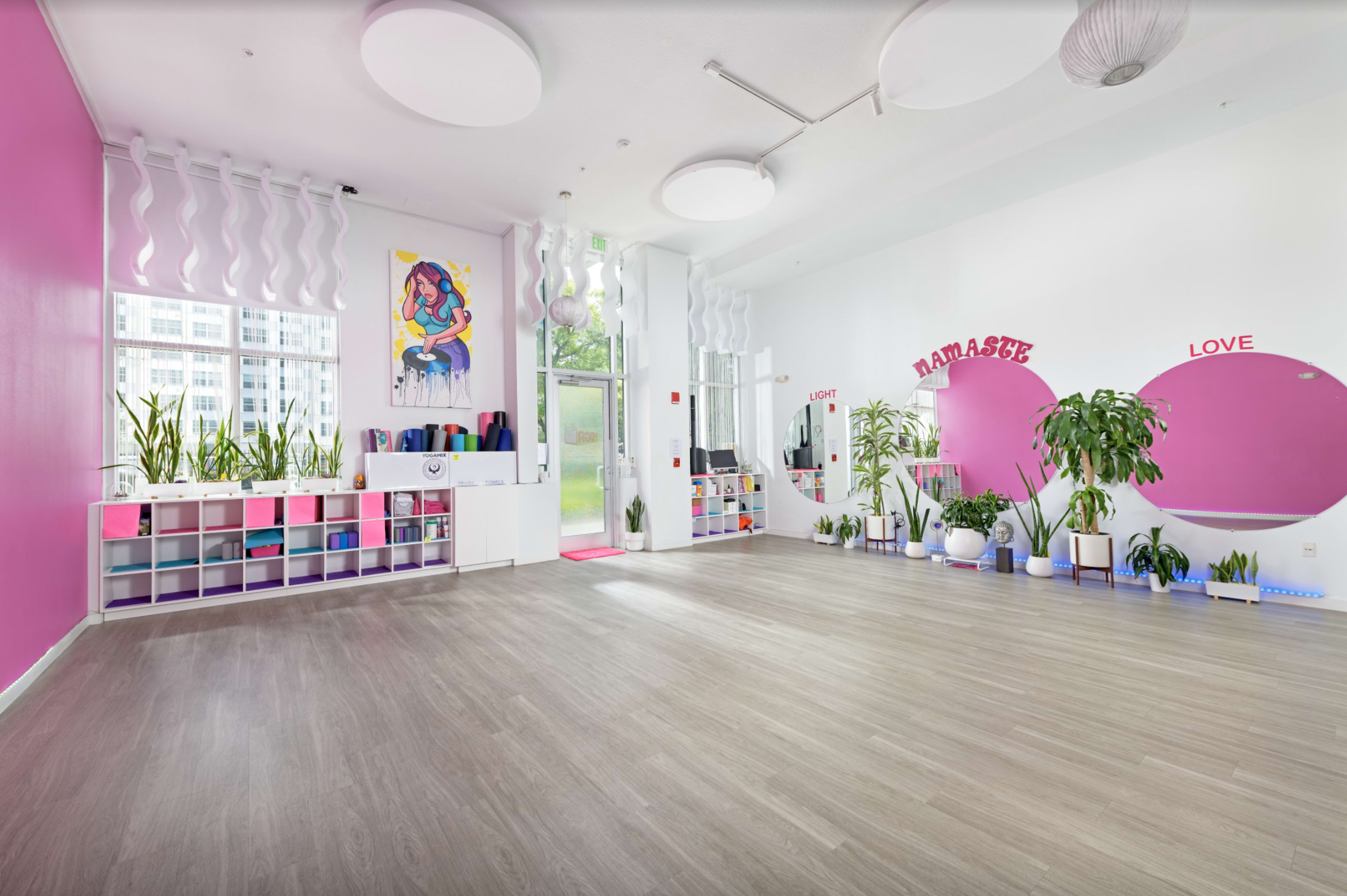 A spacious yoga studio with pink accent walls, plants, and exercise equipment neatly organized on shelves.