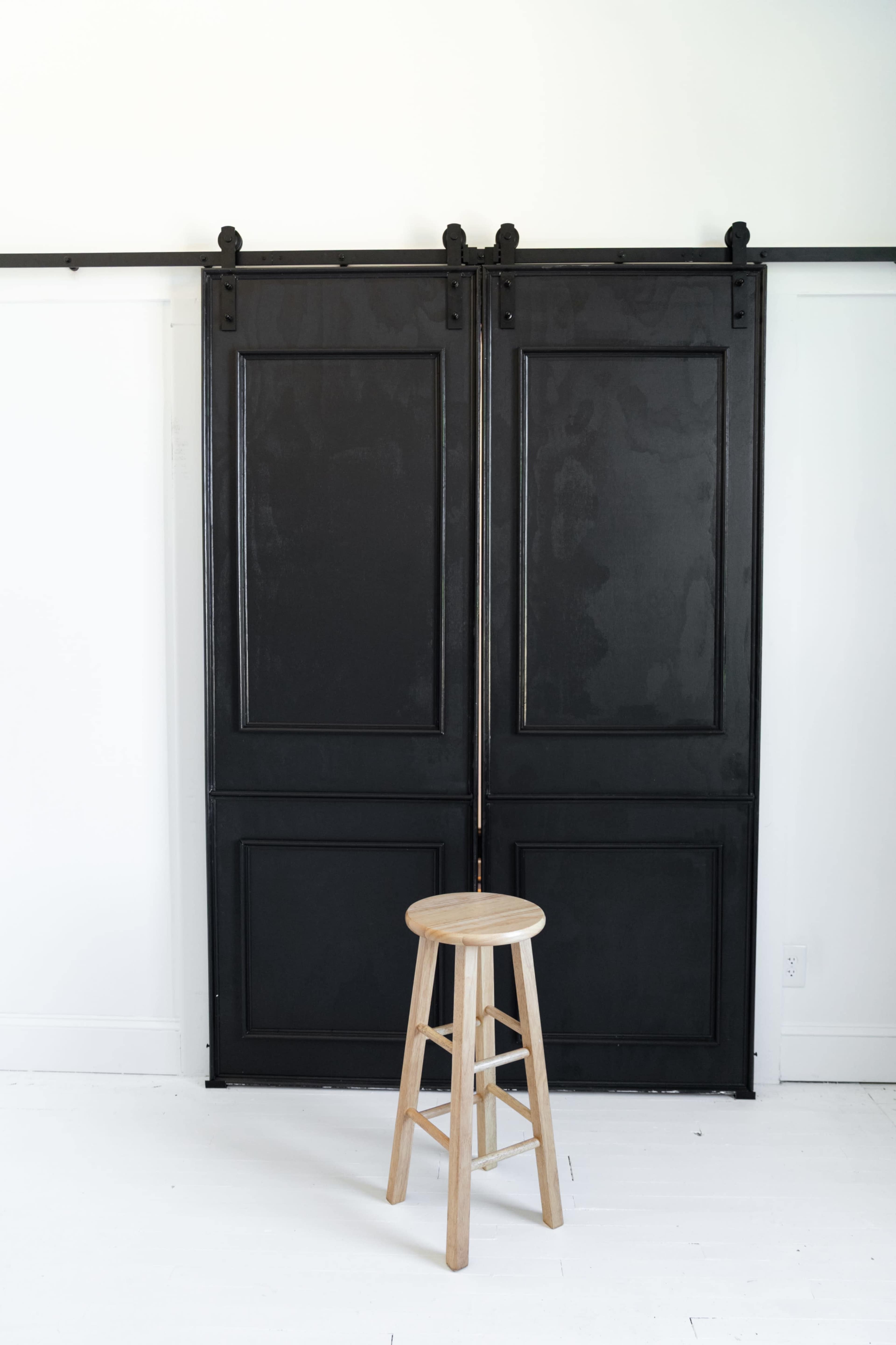 A wooden stool stands in front of a set of black sliding barn doors against a white wall.