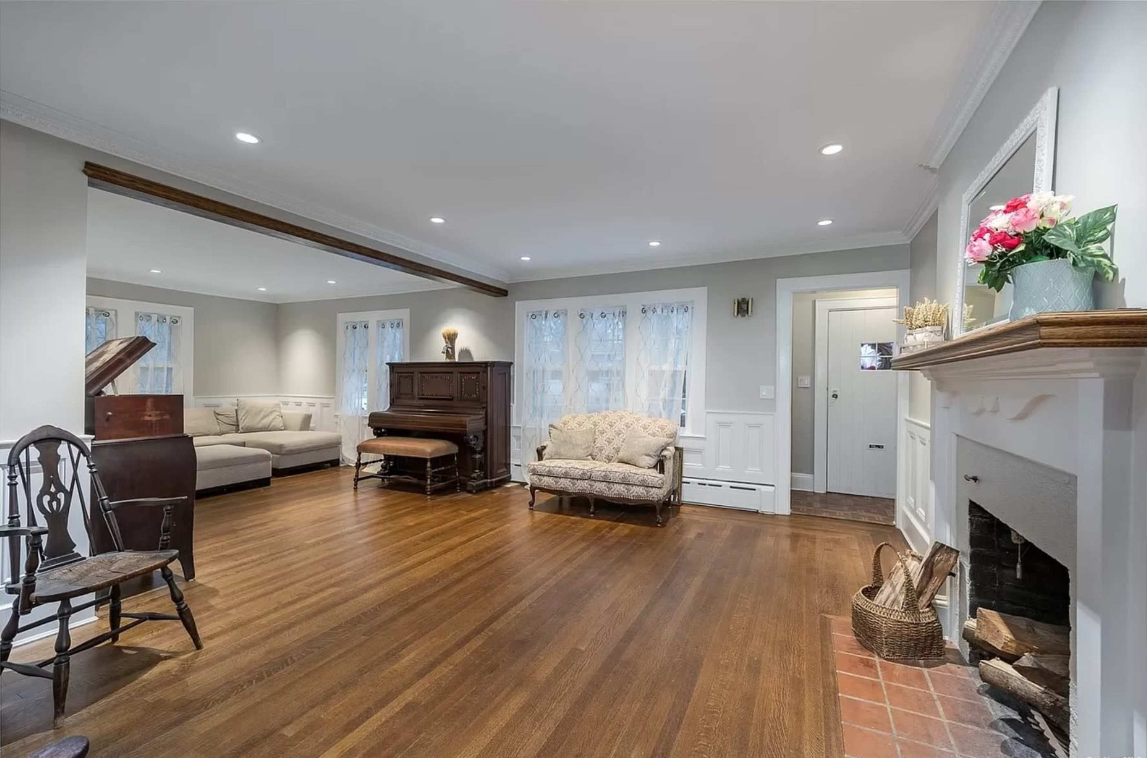 The room features hardwood floors, a vintage piano, a sofa, and a decorative fireplace with floral arrangements.