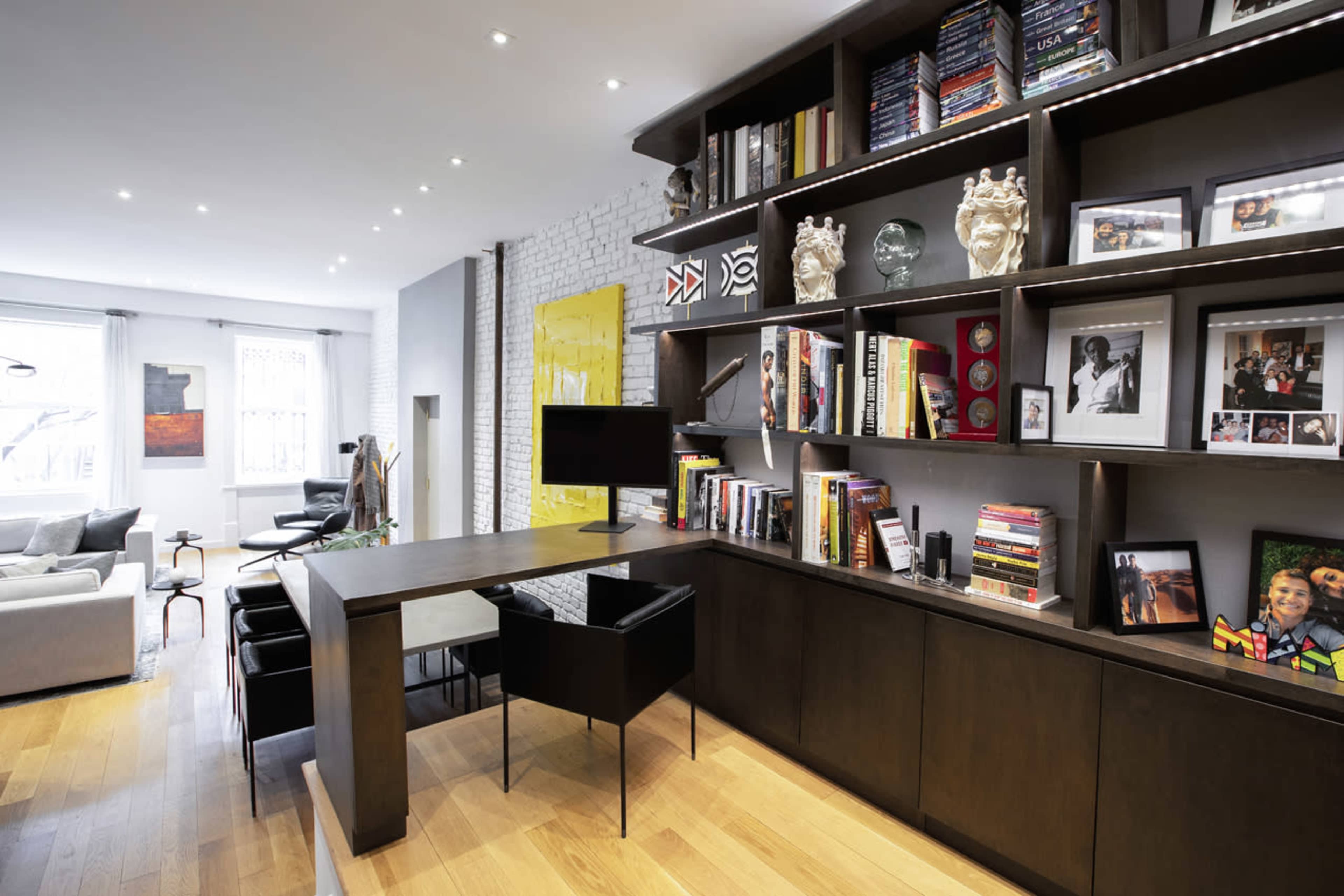 A modern home office features a long desk with a chair, a wall of built-in shelves filled with books and decorative items, and a computer monitor.