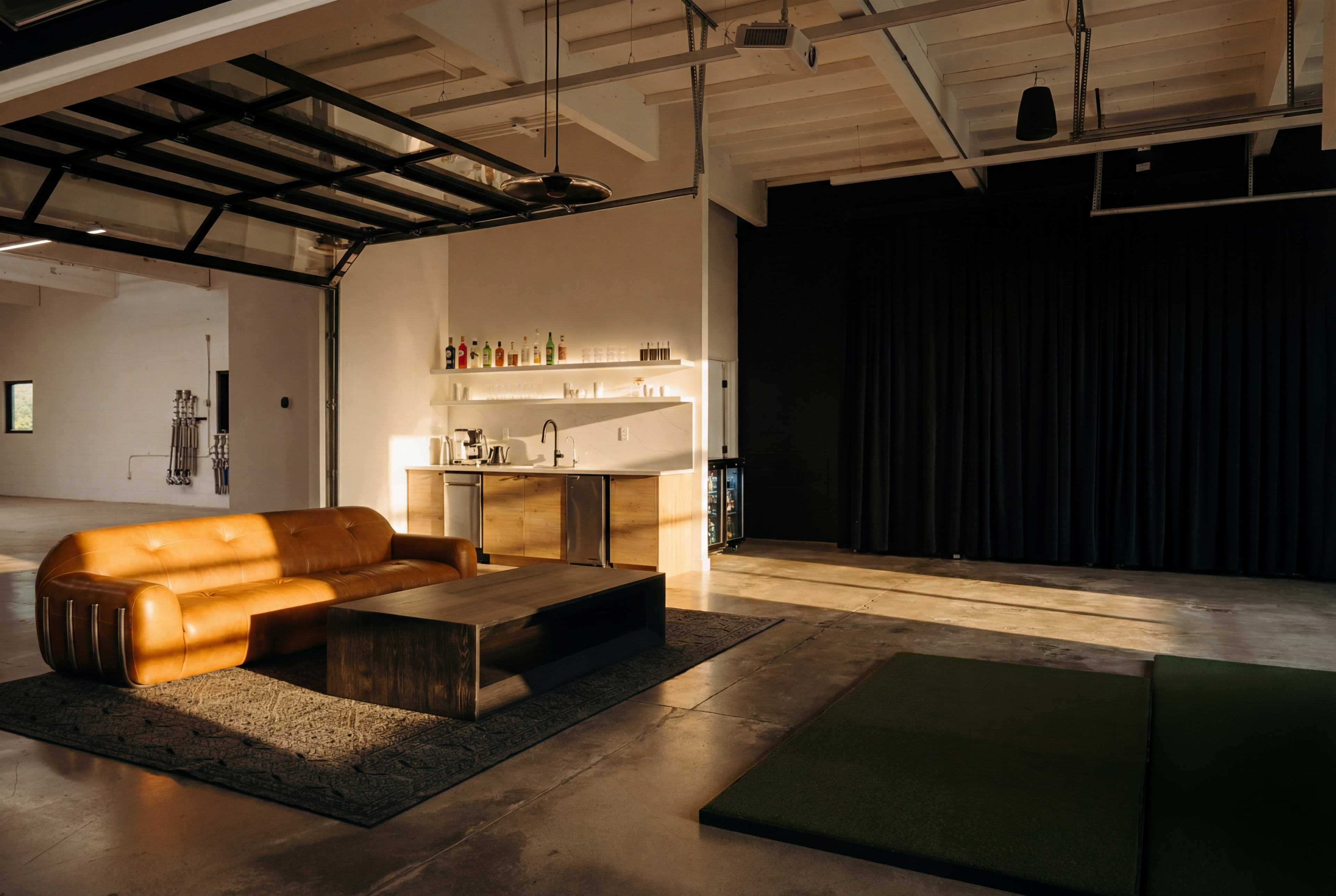 The image shows a modern open space featuring a brown leather couch, a wooden coffee table, and a kitchenette with bottles displayed on a shelf, all illuminated by natural light.