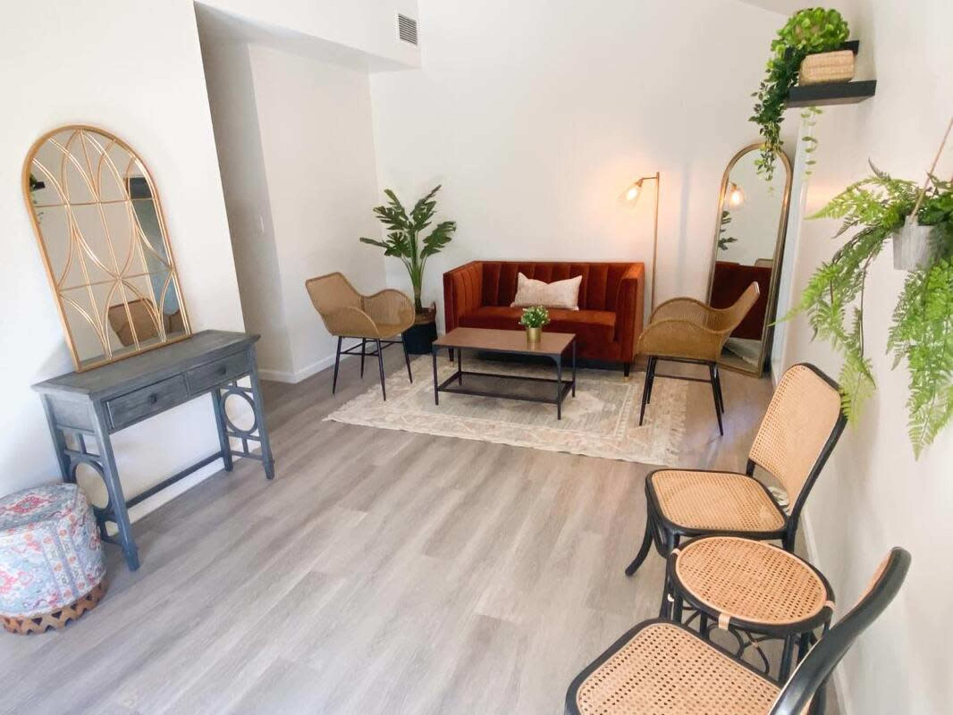 The room features a red velvet sofa, two cane chairs, a coffee table, and a floor mirror, with plants and decorative items arranged throughout.