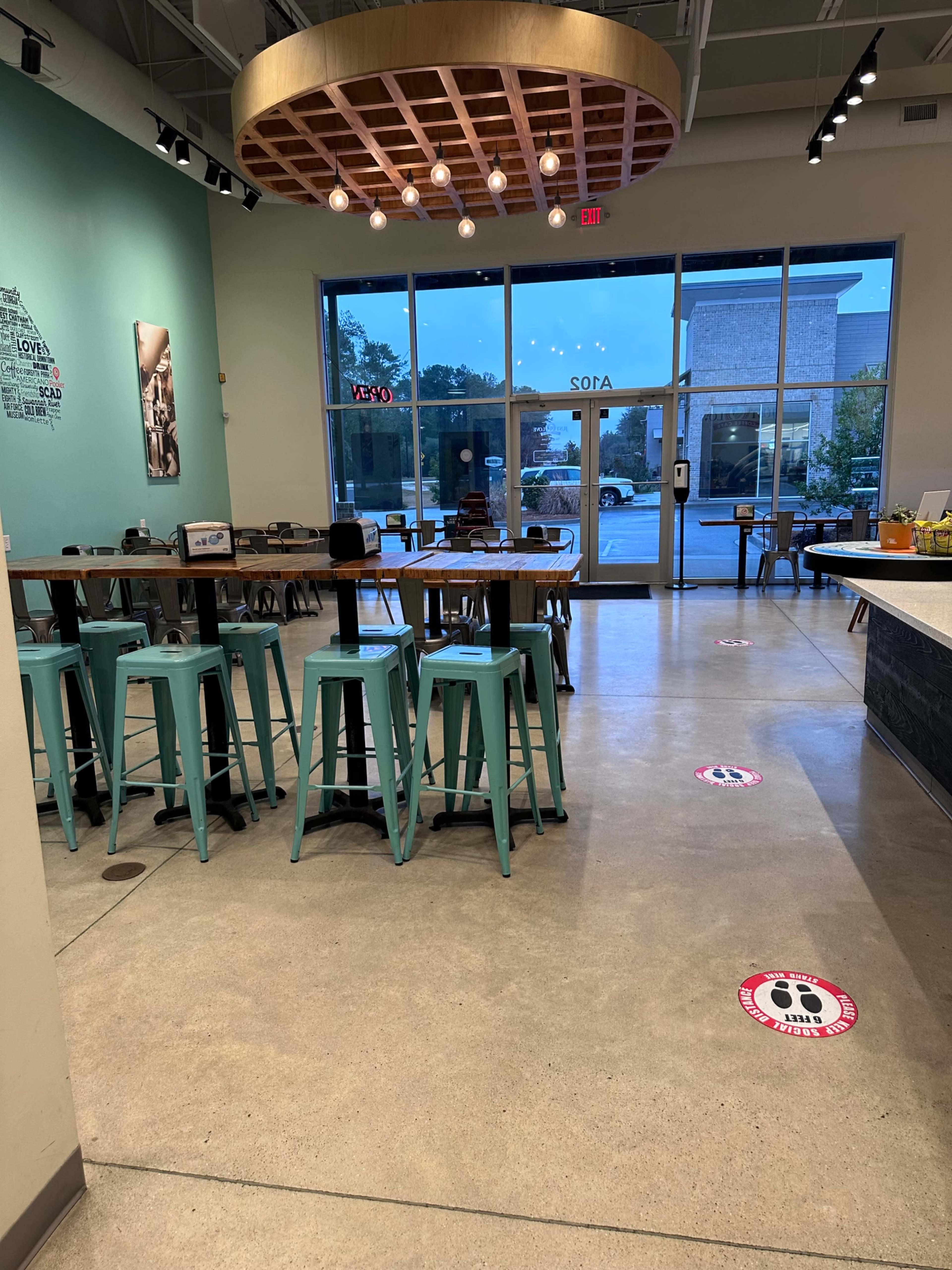 A modern café interior with turquoise stools and tables, illuminated by overhead lights, and social distancing markers on the floor.