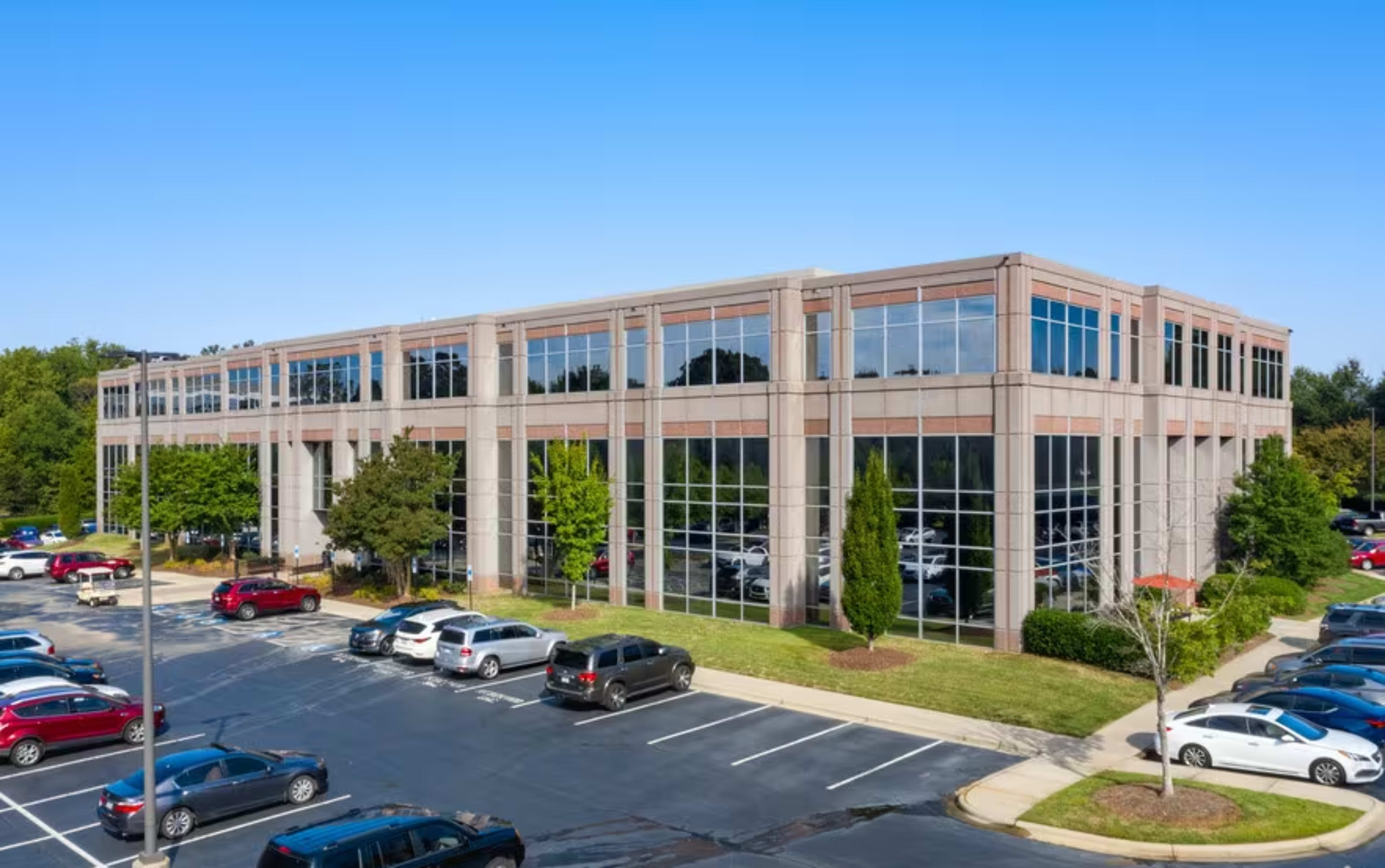 A modern office building with large windows surrounded by landscaped trees and a parking lot filled with vehicles.