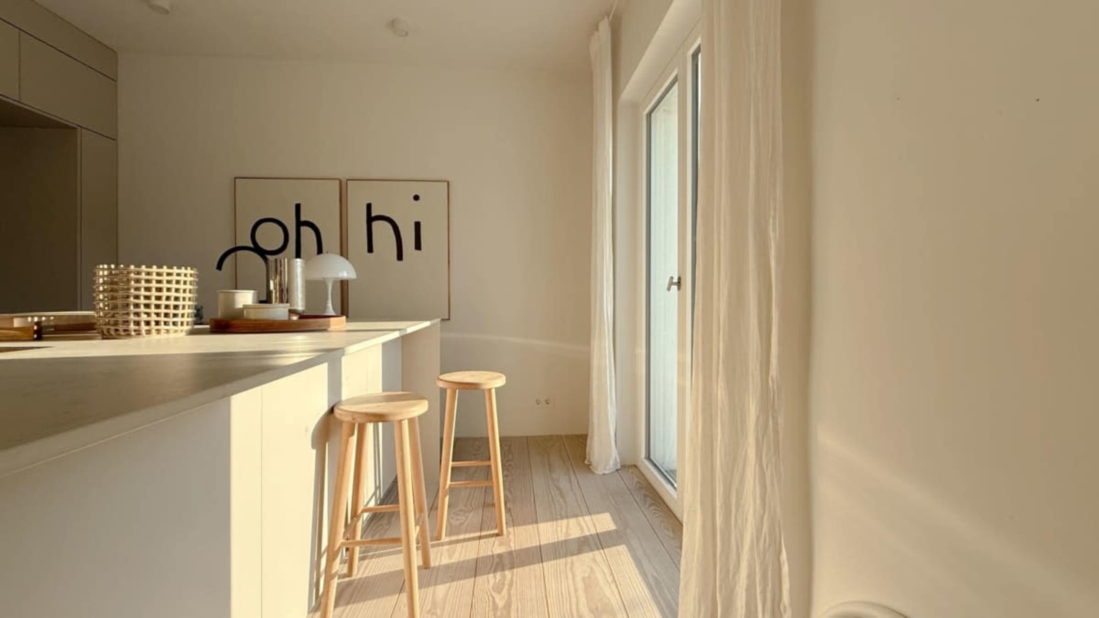 The image shows a minimalist kitchen space featuring a light-colored countertop with two wooden stools, large windows allowing natural light in, and two wall art pieces displaying the words "oh" and "hi."