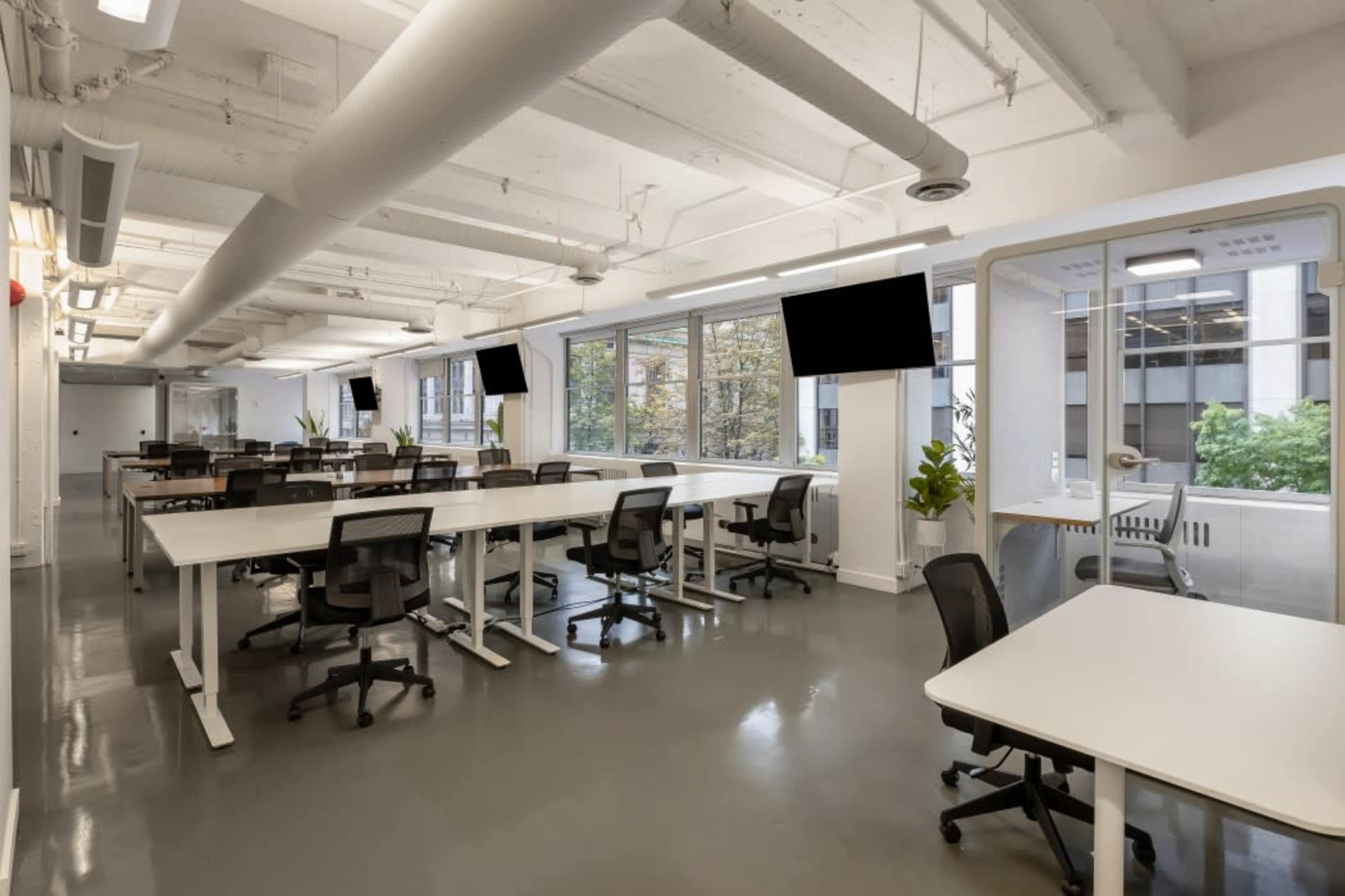 A modern office space features multiple adjustable desks and chairs, large windows, and two mounted televisions.