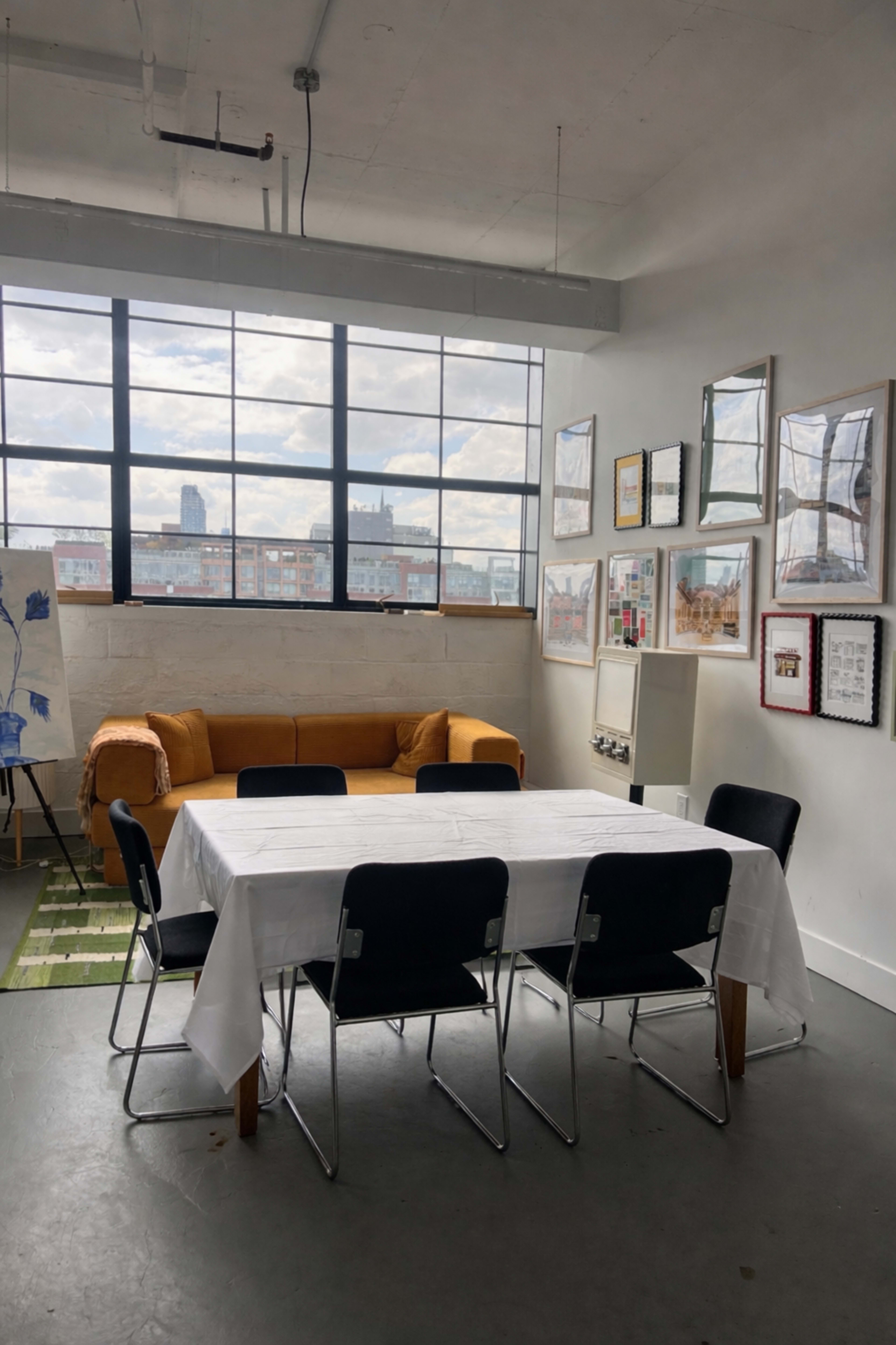 The image shows a bright room featuring a yellow sofa, a large white table surrounded by black chairs, and several framed artworks on the walls next to a large window overlooking a cityscape.