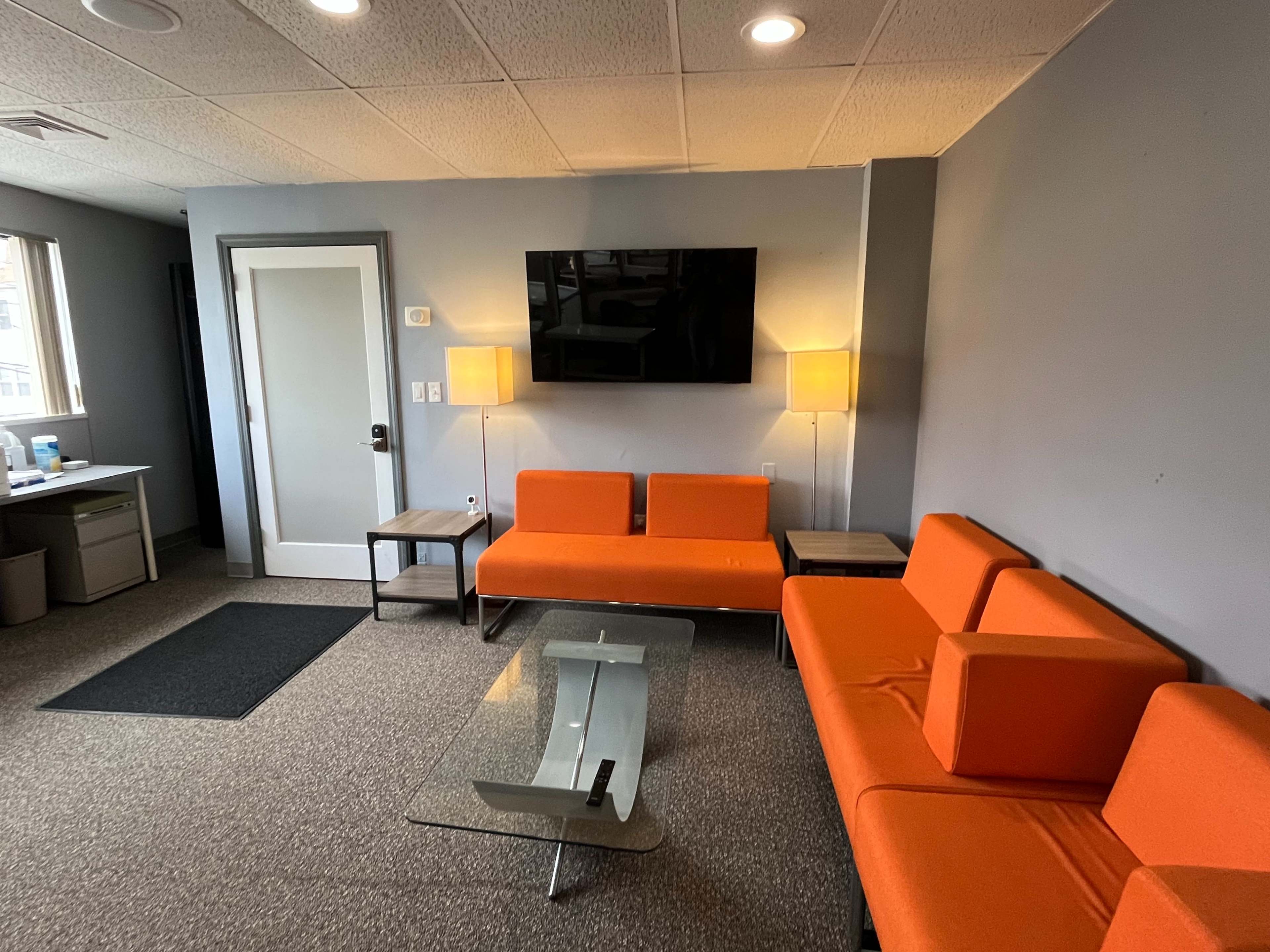The image shows a modern lounge area with two orange couches, a glass coffee table, and a television mounted on the wall.