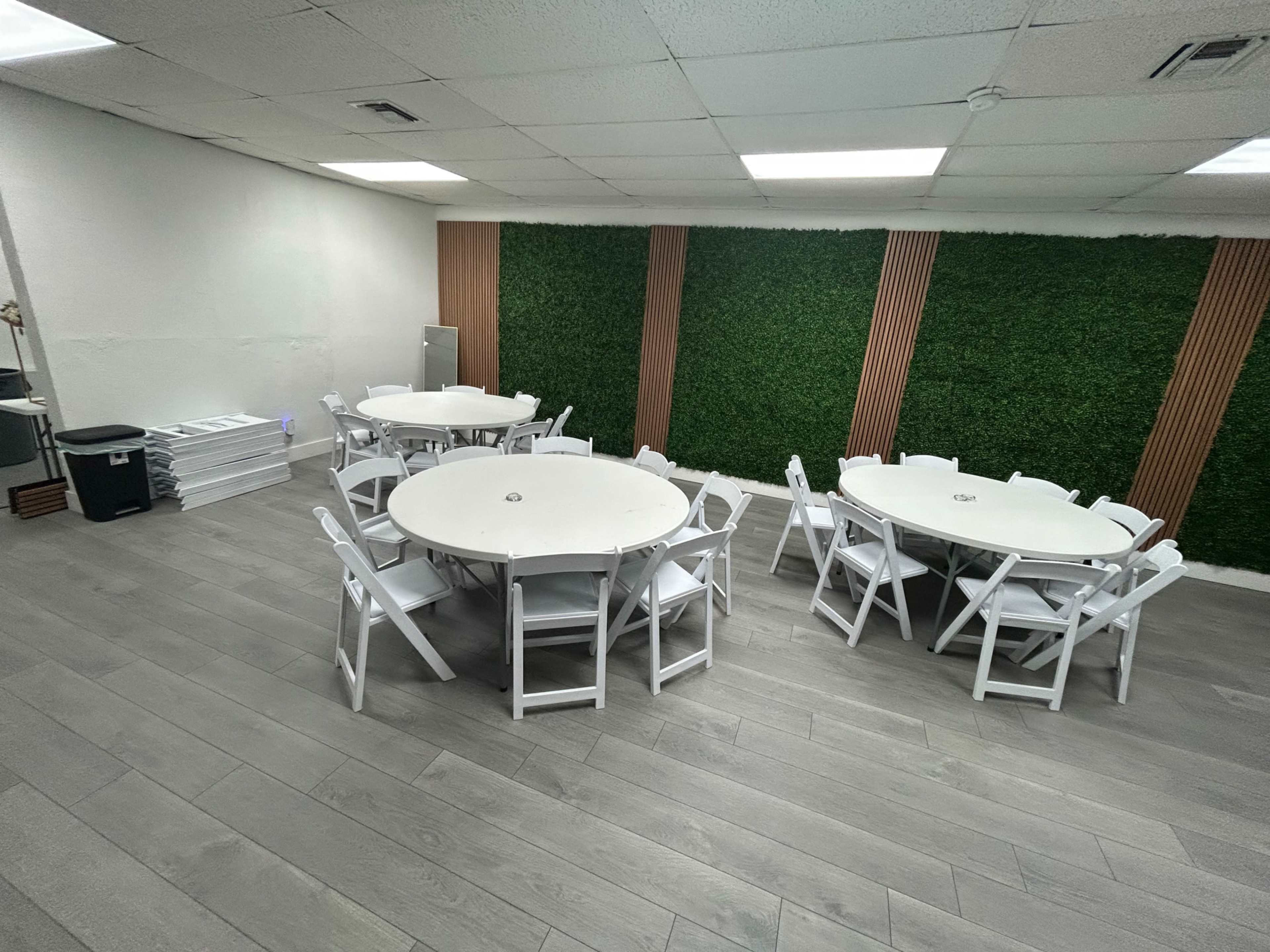 The image shows a spacious room with several round tables surrounded by white chairs, and a green wall feature with wooden accents.