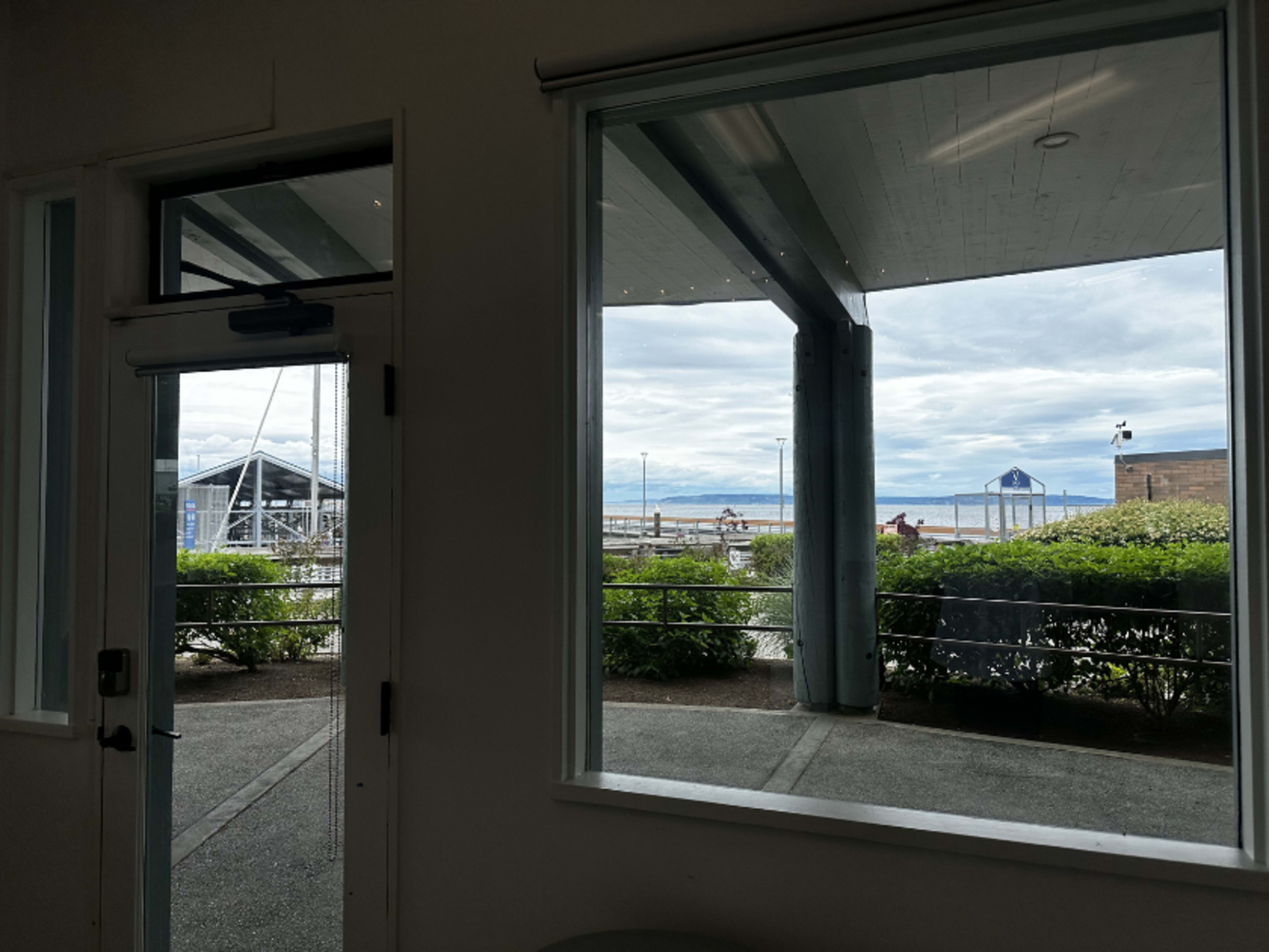 The image shows a view from inside a building, looking out through large windows towards a dock and waterfront area under a cloudy sky.