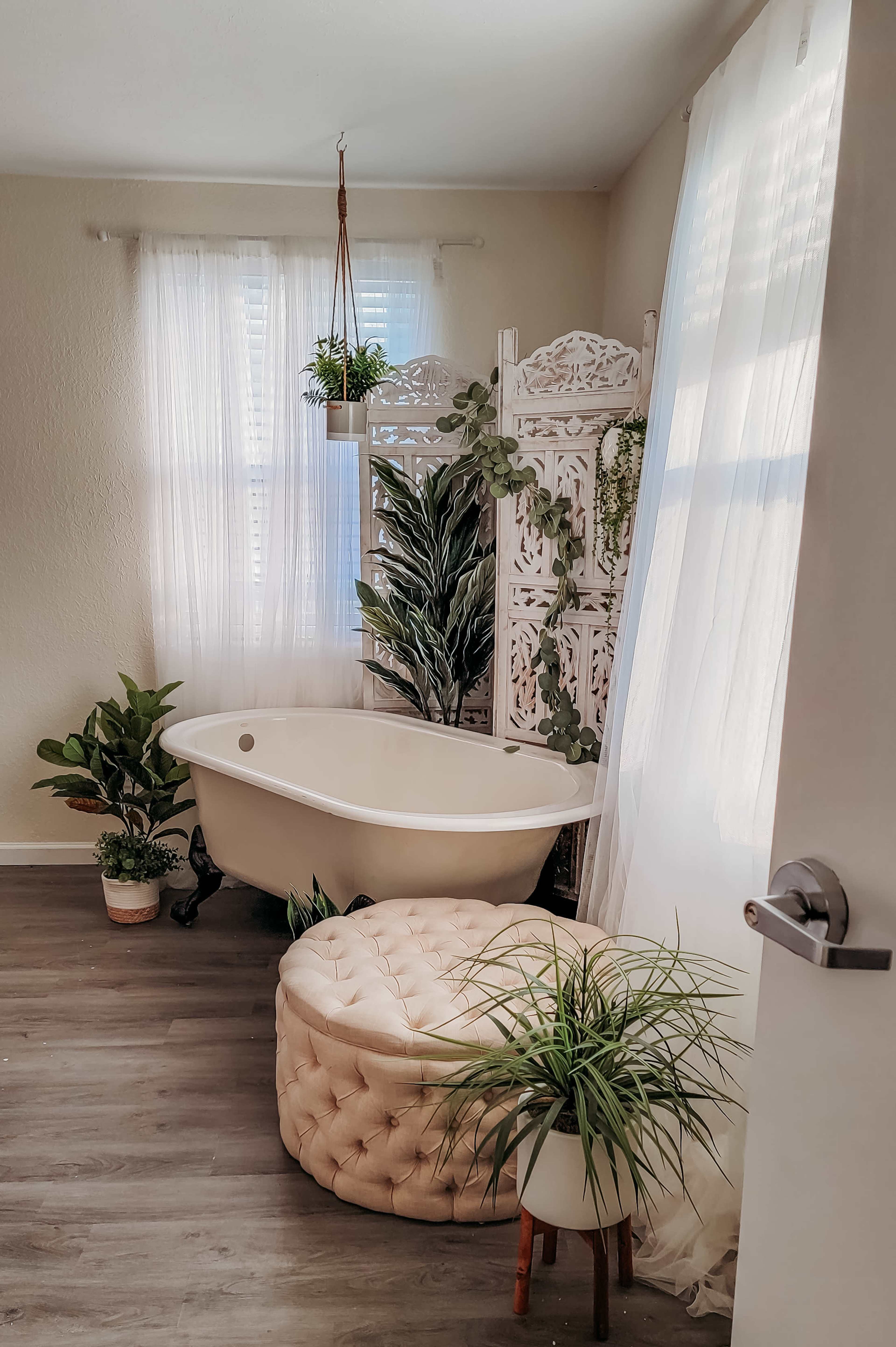 A bright bathroom featuring a freestanding bathtub, a large potted plant, a decorative wooden screen, and a tufted ottoman near a window.