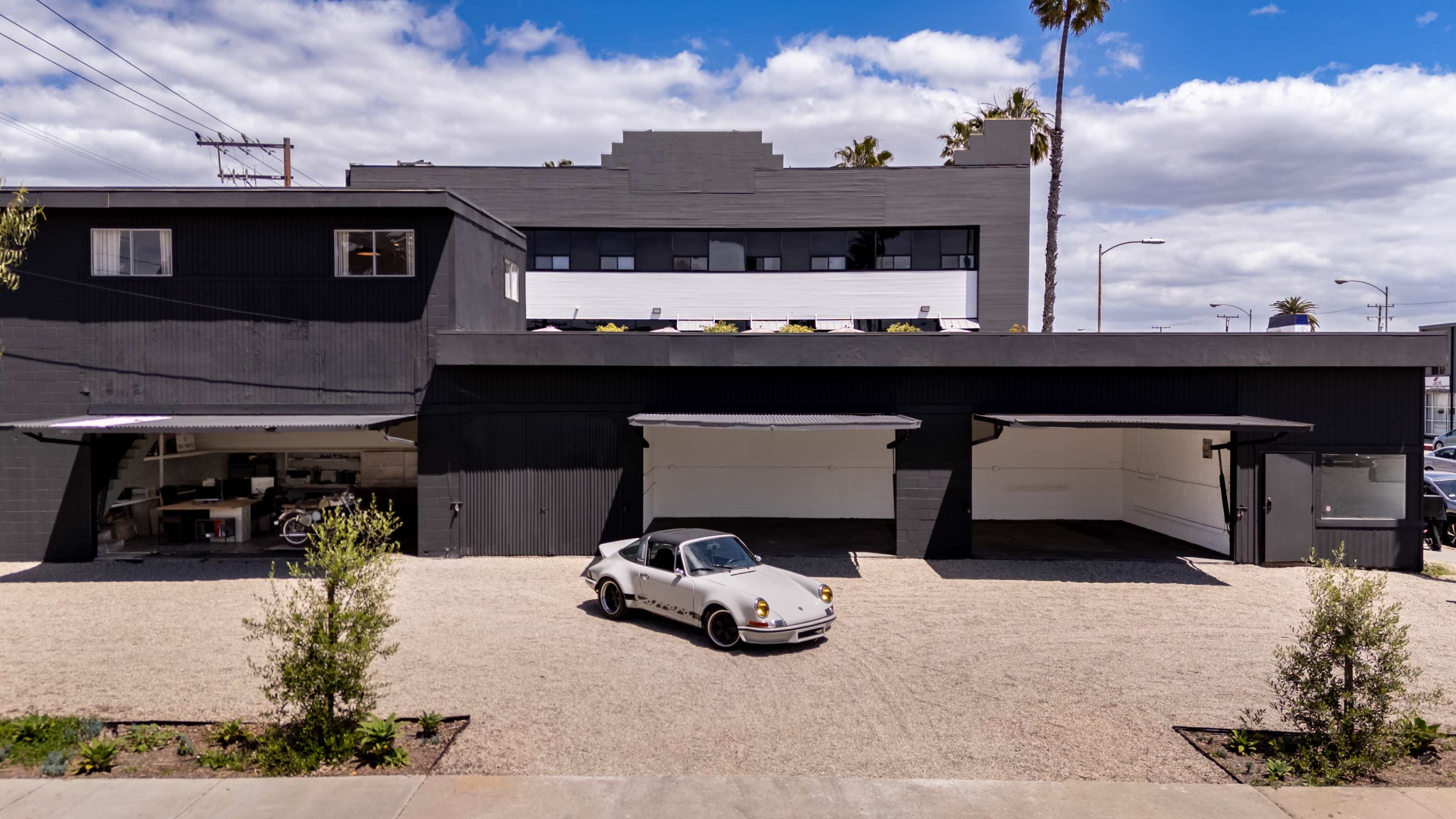 A vintage car is parked in front of a black building with multiple garage doors and a palm tree in view under a cloudy sky.