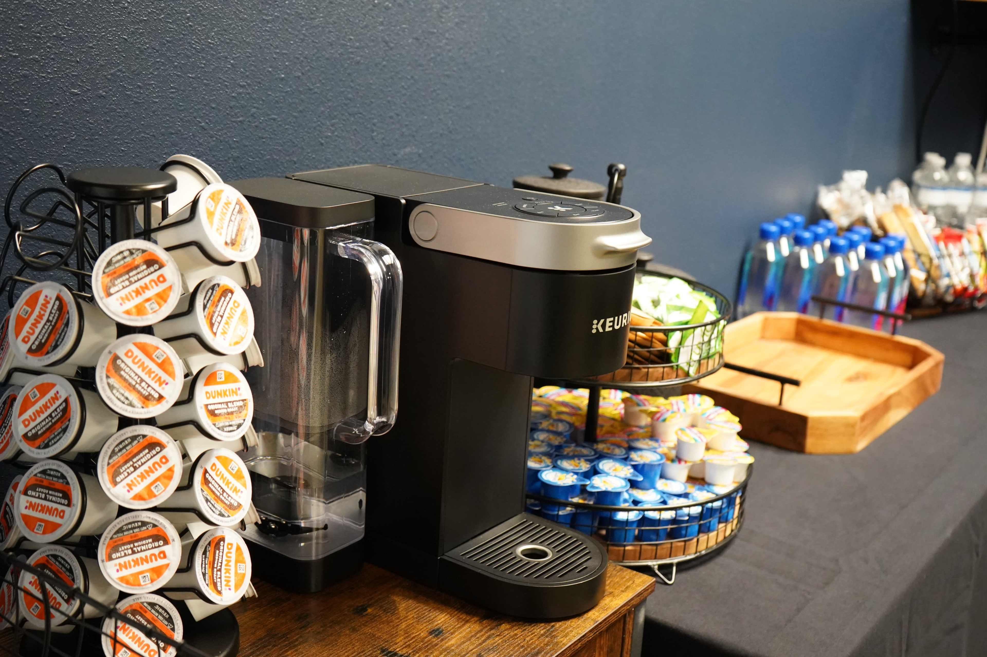 A coffee maker alongside a selection of coffee pods and creamers on a table with water bottles and snacks in the background.