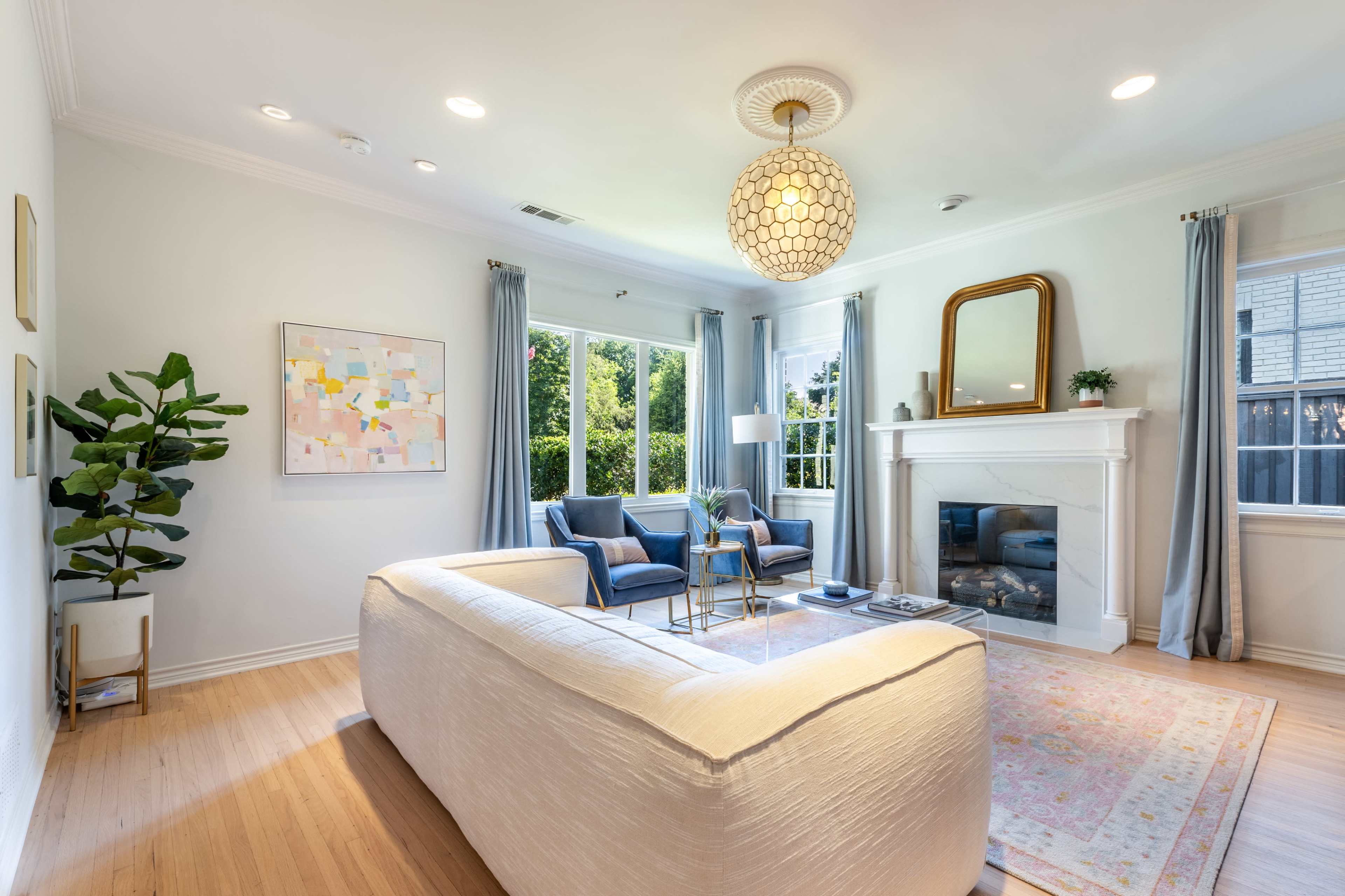A cozy living room features a light-colored sofa, two blue chairs, a coffee table, and a fireplace with a mirror above it, surrounded by large windows and a potted plant.
