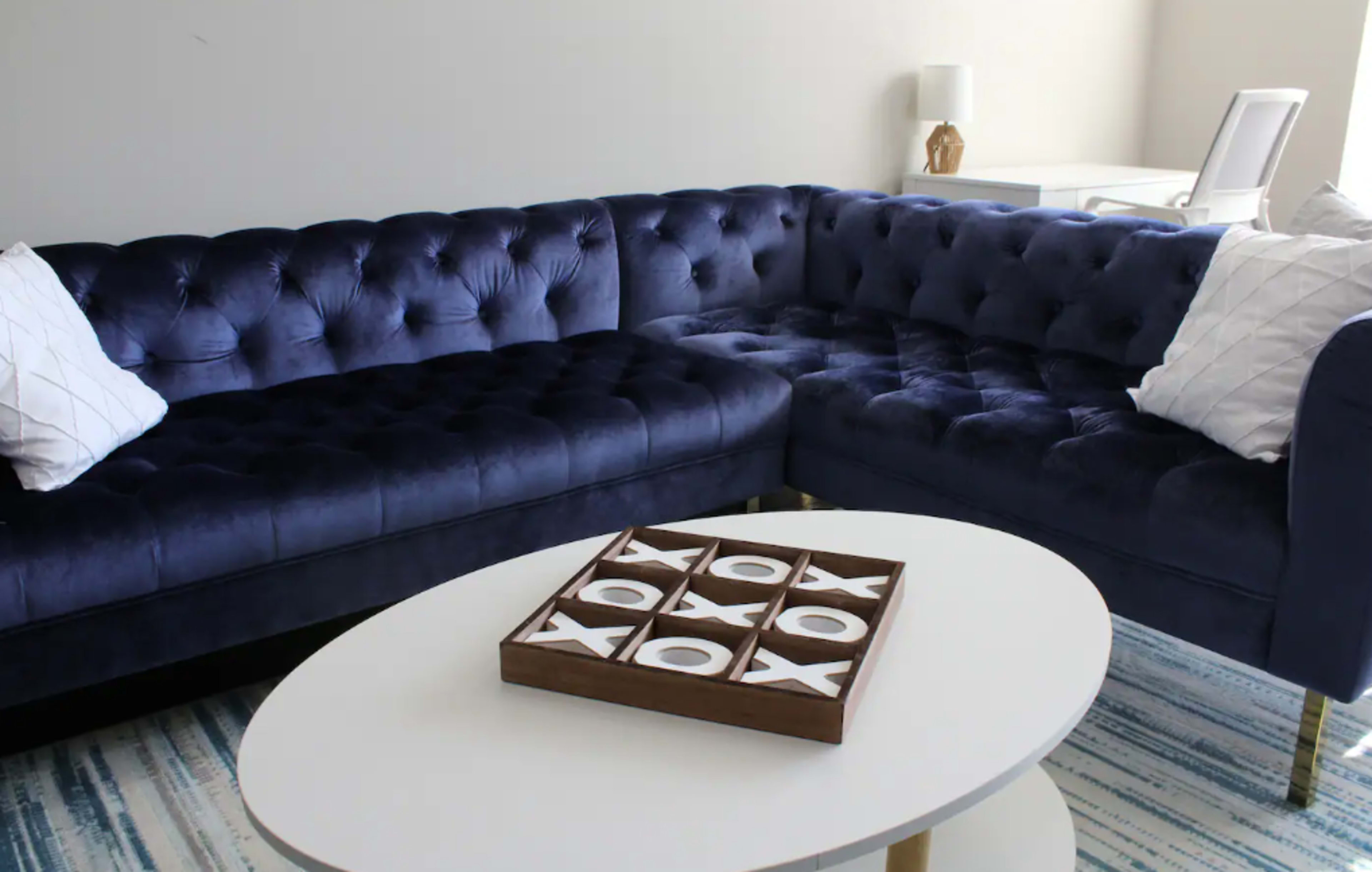 A navy blue velvet sectional sofa with tufted upholstery is positioned next to a round white coffee table featuring a tic-tac-toe design.