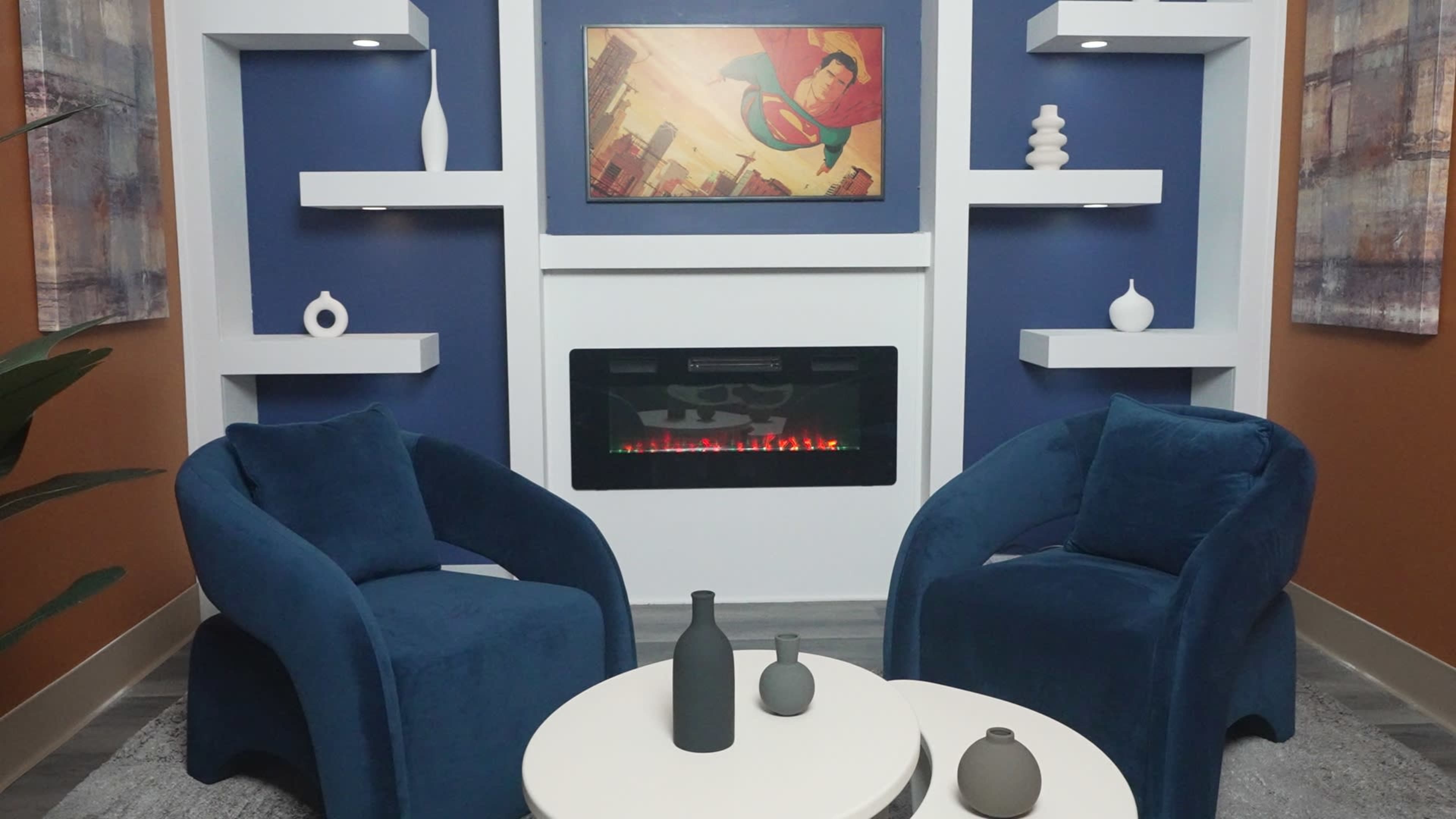 The image shows a contemporary living area with two blue upholstered chairs facing a small circular table, flanked by a wall-mounted fireplace and a framed superhero artwork.