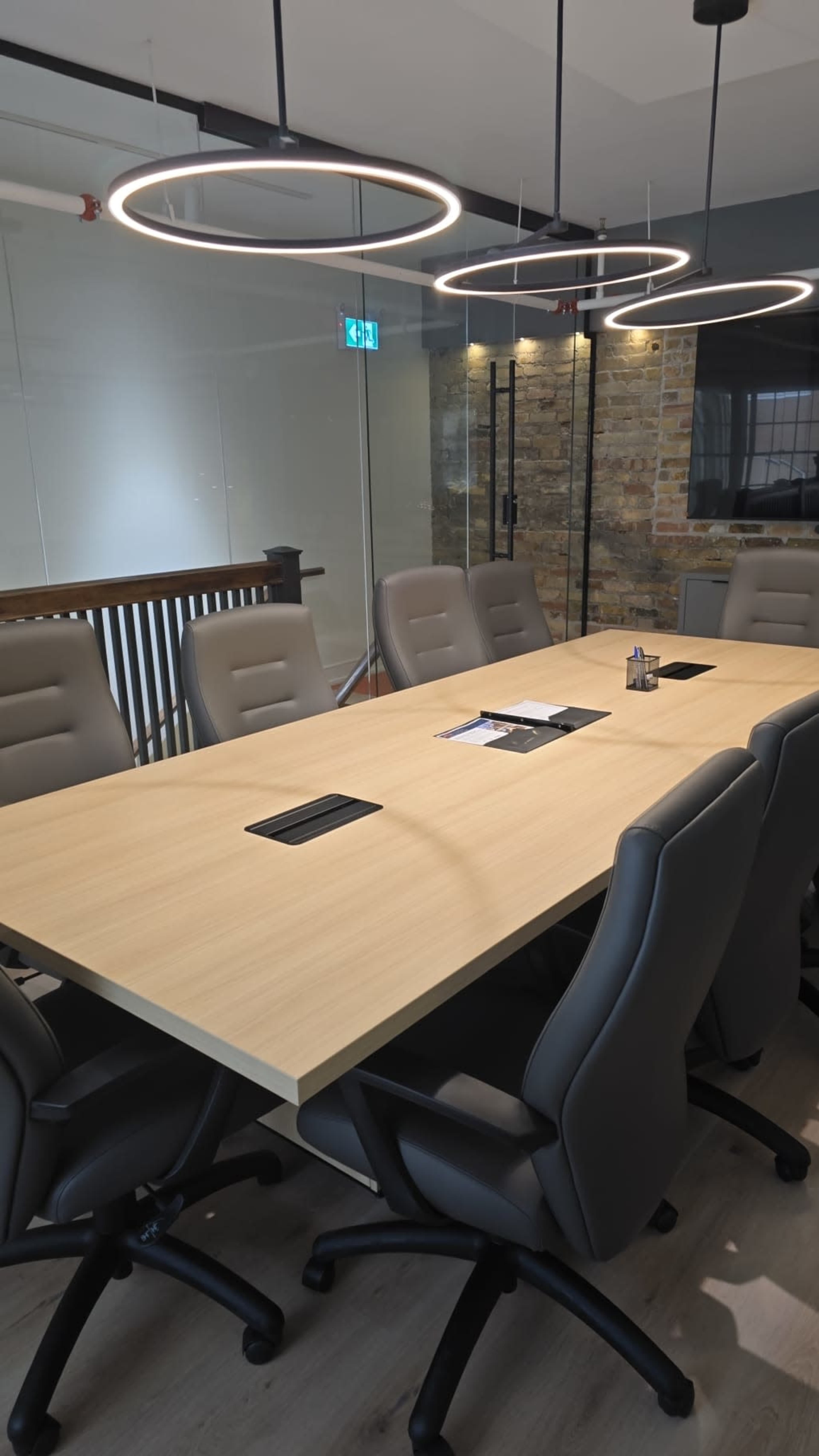 A modern conference room features a long wooden table surrounded by ergonomic chairs and illuminated by circular hanging LED lights.