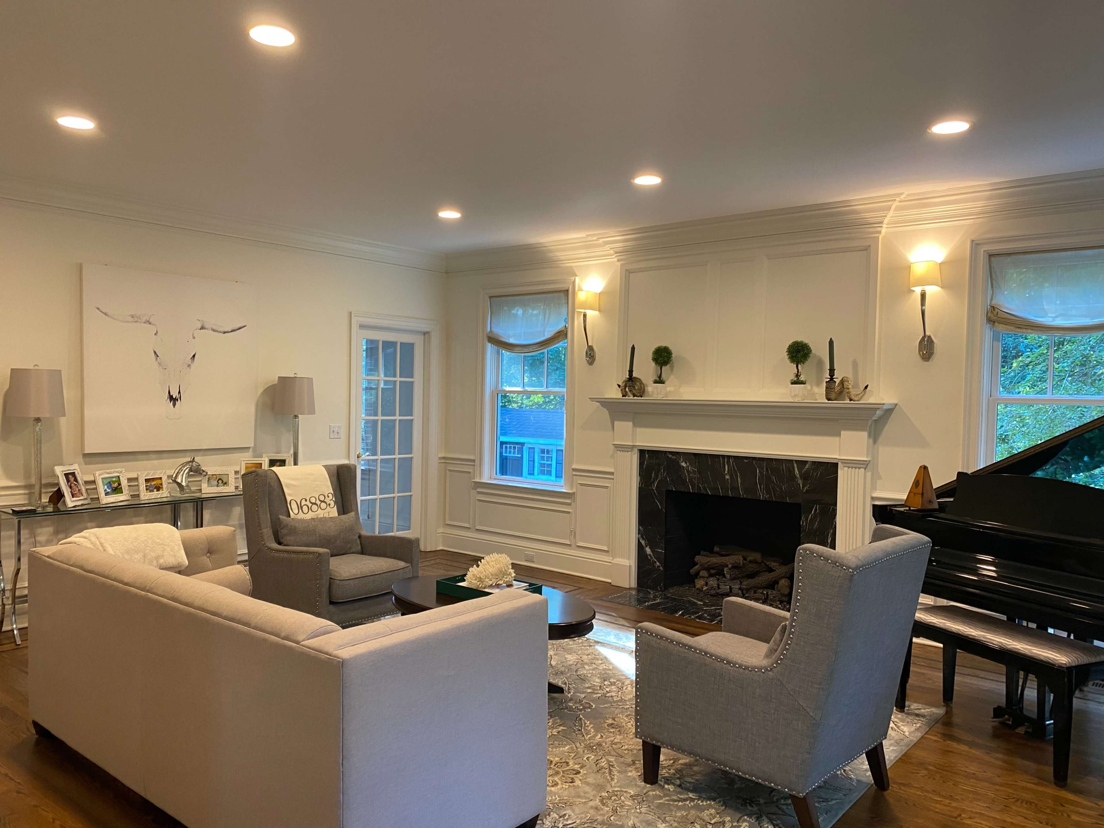 The living room features a large sofa, two armchairs, a coffee table, a black piano, and a fireplace, with a mix of natural light and overhead lighting illuminating the space.