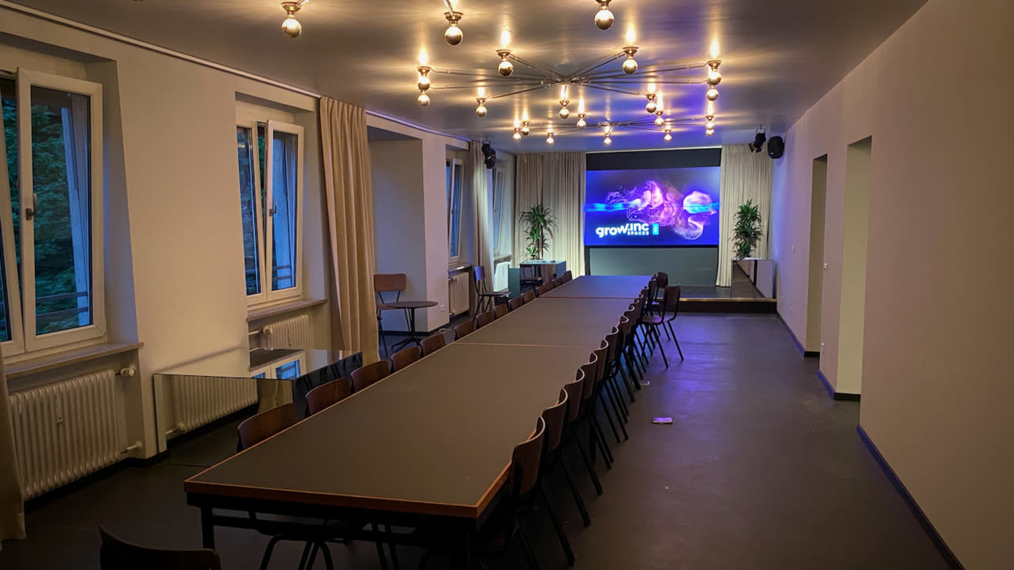 A large conference room features a long table with chairs arranged around it, a screen displaying "growing" on the wall, and multiple light fixtures overhead.