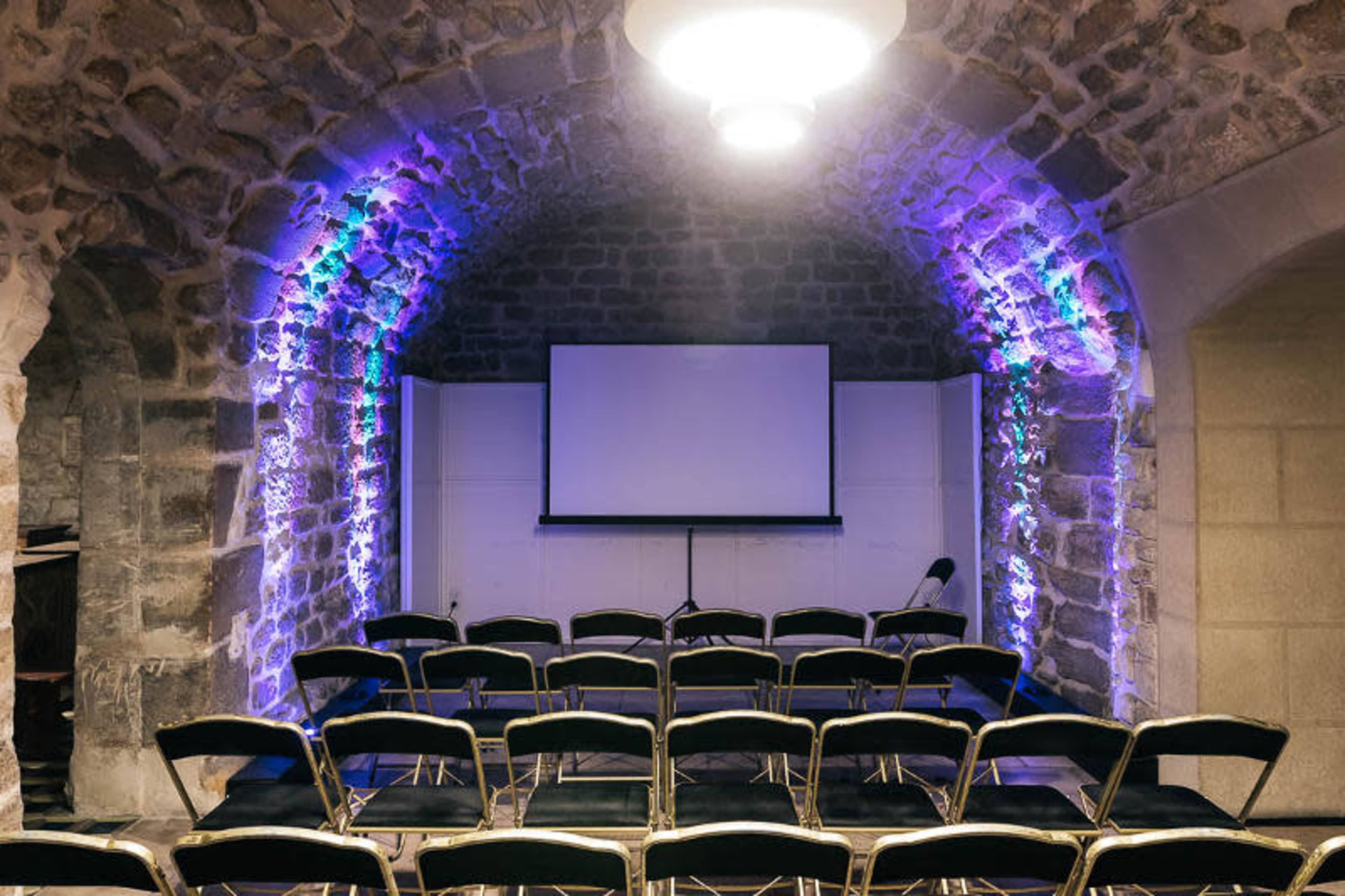 A stone-walled meeting room features rows of black chairs facing a large screen, illuminated by colored lights.